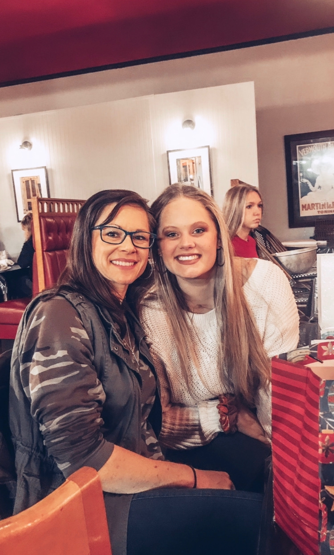 Two people pose together at a restaurant table, with framed pictures and other diners in the background.