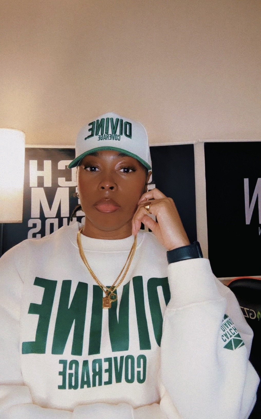 Woman wearing a white cap and sweatshirt with green text, gold necklace, and ring, indoors with black and white background.