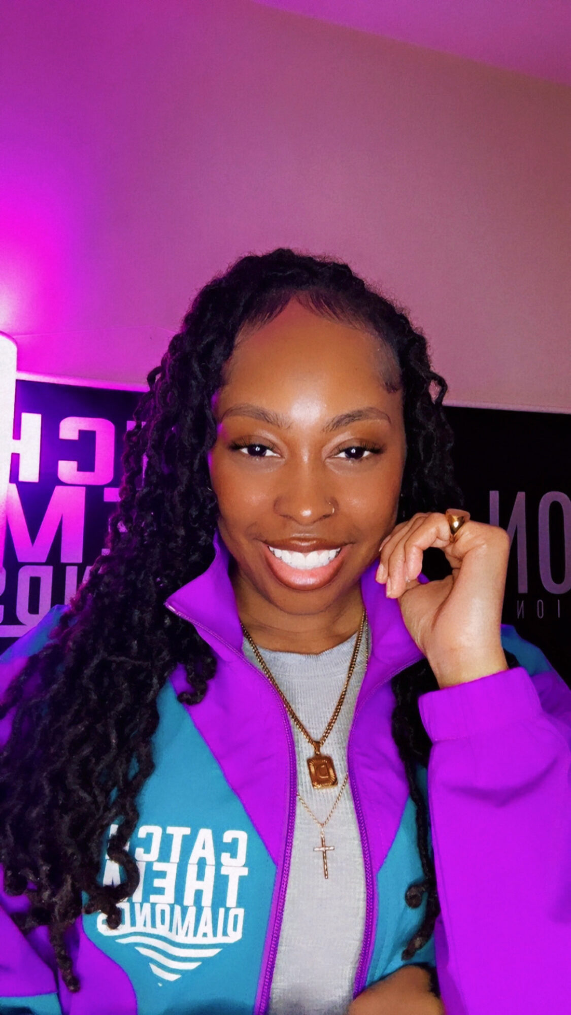 Smiling woman with long curly hair wearing a purple jacket and gray shirt, holding her collar, in a room with purple lighting.