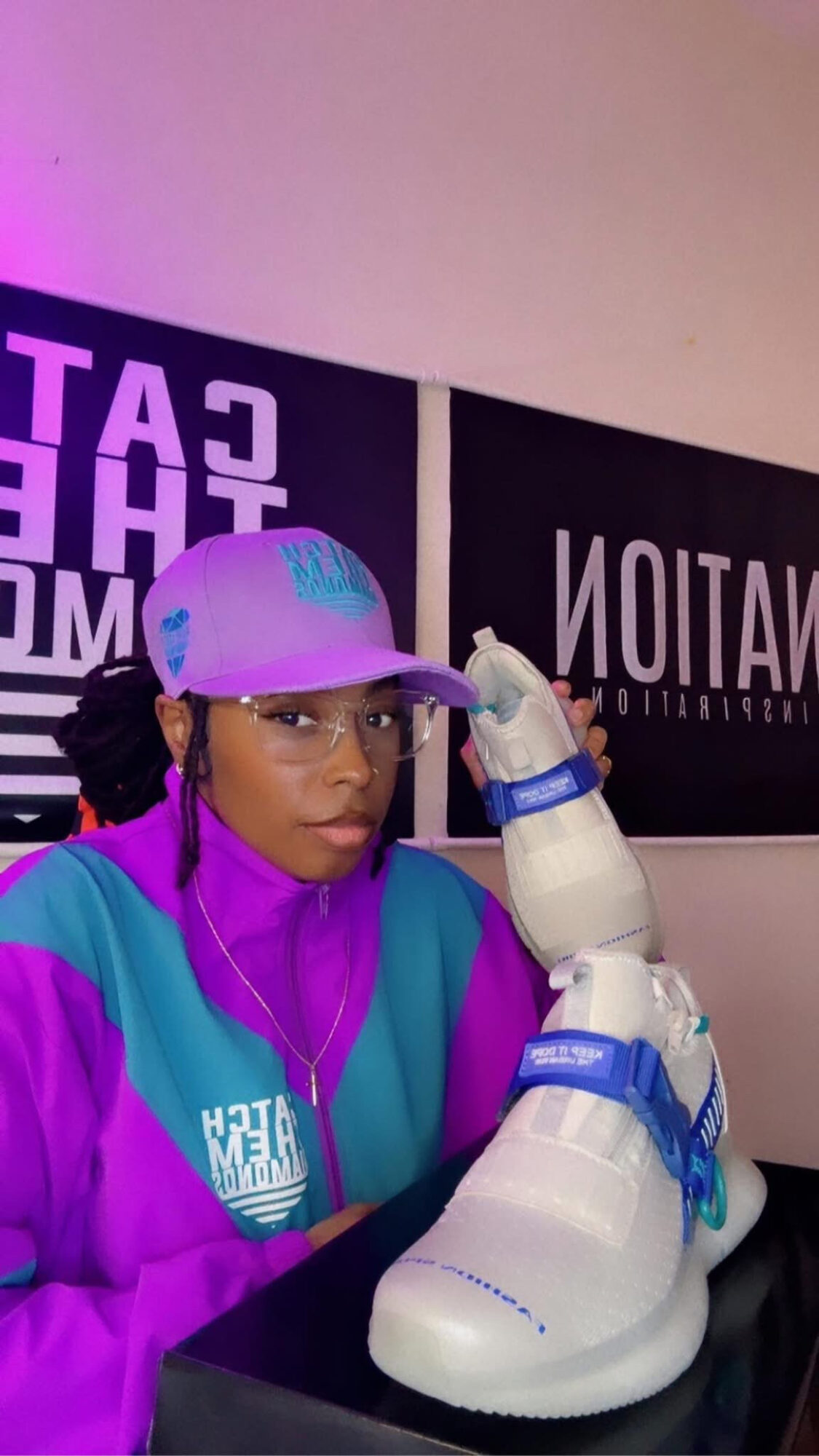 Person wearing a purple and teal jacket and cap, with a robotic arm and a large white shoe, in front of a black sign.