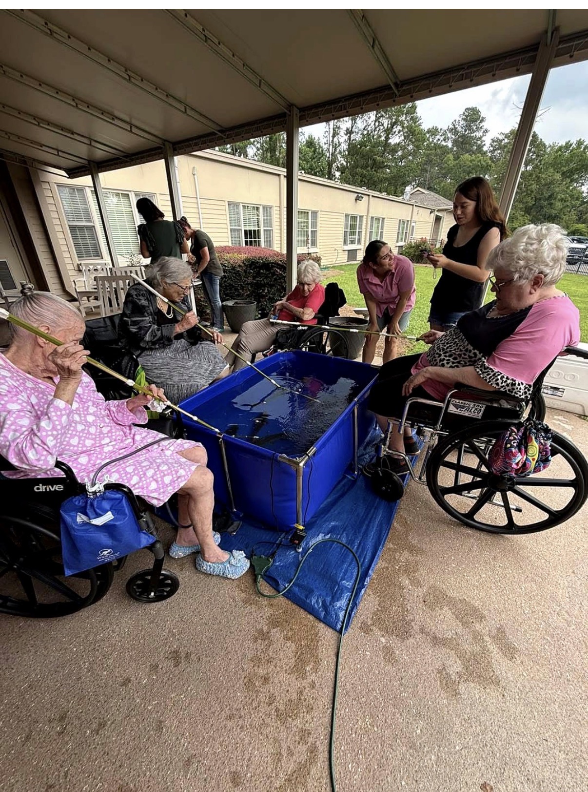 Group of elderly and young people gathered outdoors under a covered area, some in wheelchairs, around a blue water activity setup.
