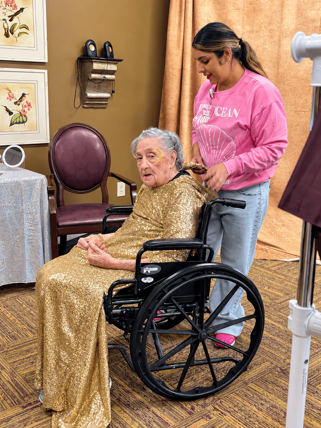 A woman in pink assists an elderly woman in a wheelchair in a room with artwork and furniture.