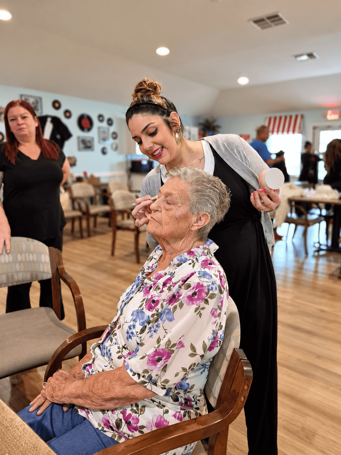 Young woman smiling and leaning toward elderly woman seated in a chair, in a room with other people and decorations.