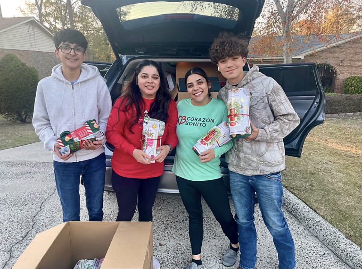 Four people stand outdoors holding wrapped gifts, with a car and trees in the background.