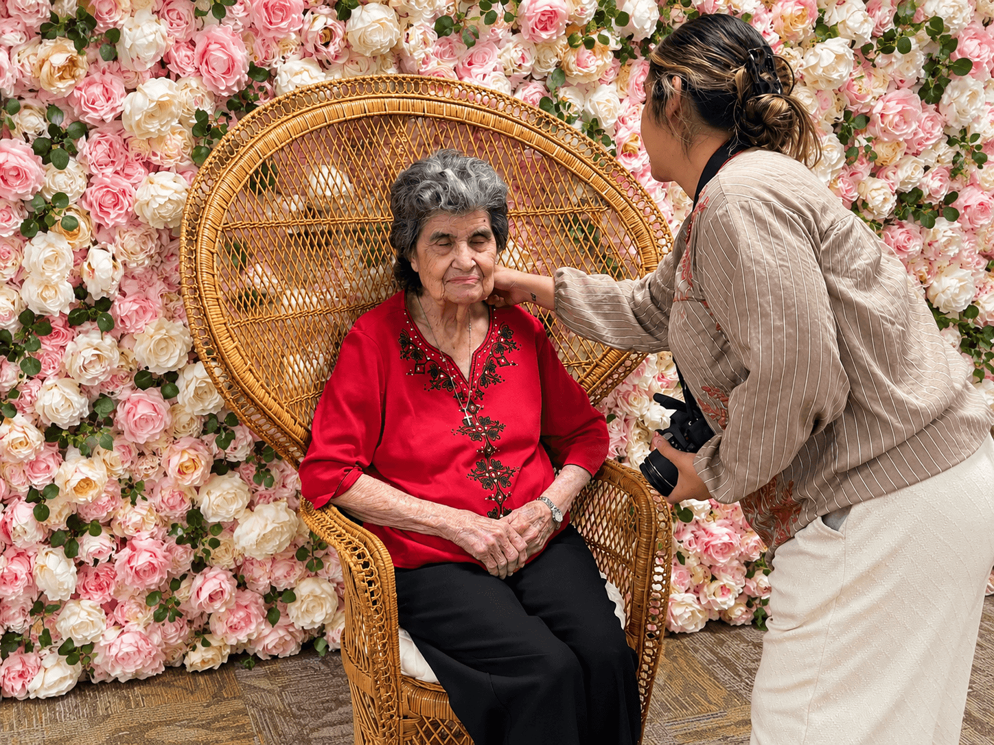 A woman in a beige jacket and white pants touches an elderly woman in a red blouse sitting on a wicker chair, with a floral backdrop.