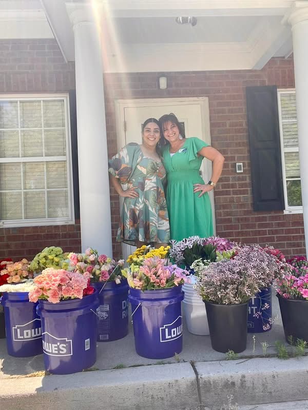 Two women standing in front of a brick house with flower buckets in the foreground.