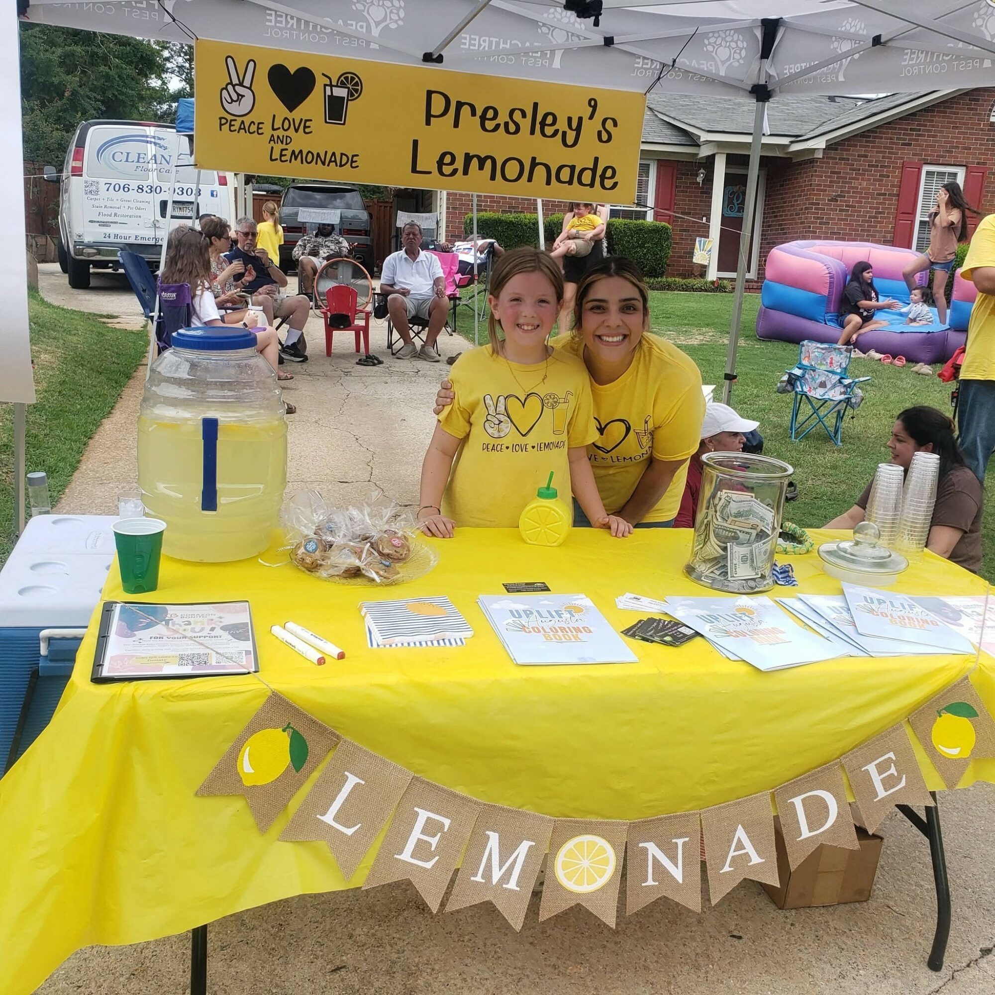 Two smiling people behind a lemonade stand with a yellow tablecloth and a banner reading 'Presley's Lemonade.'
