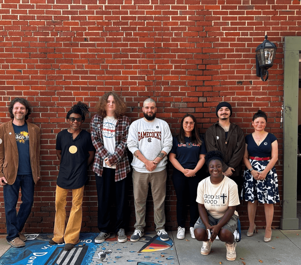 Group of seven diverse people standing against a brick wall outdoors, smiling, with a sidewalk in front.