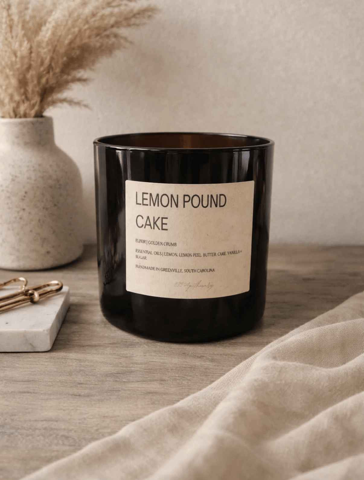 Black candle with a label reading 'LEMON POUND CAKE' on a wooden surface, with a beige cloth and a vase with dried plants nearby.