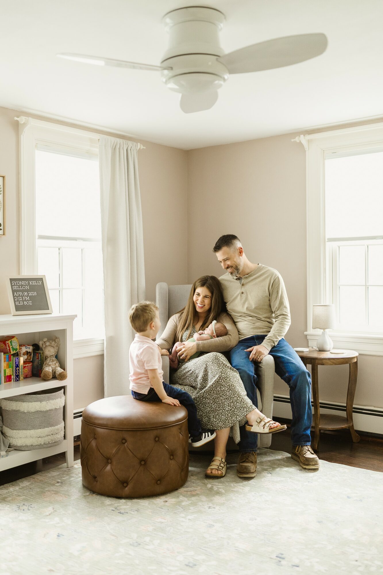 Family sitting on a brown ottoman in a bright living room with large windows and white walls.