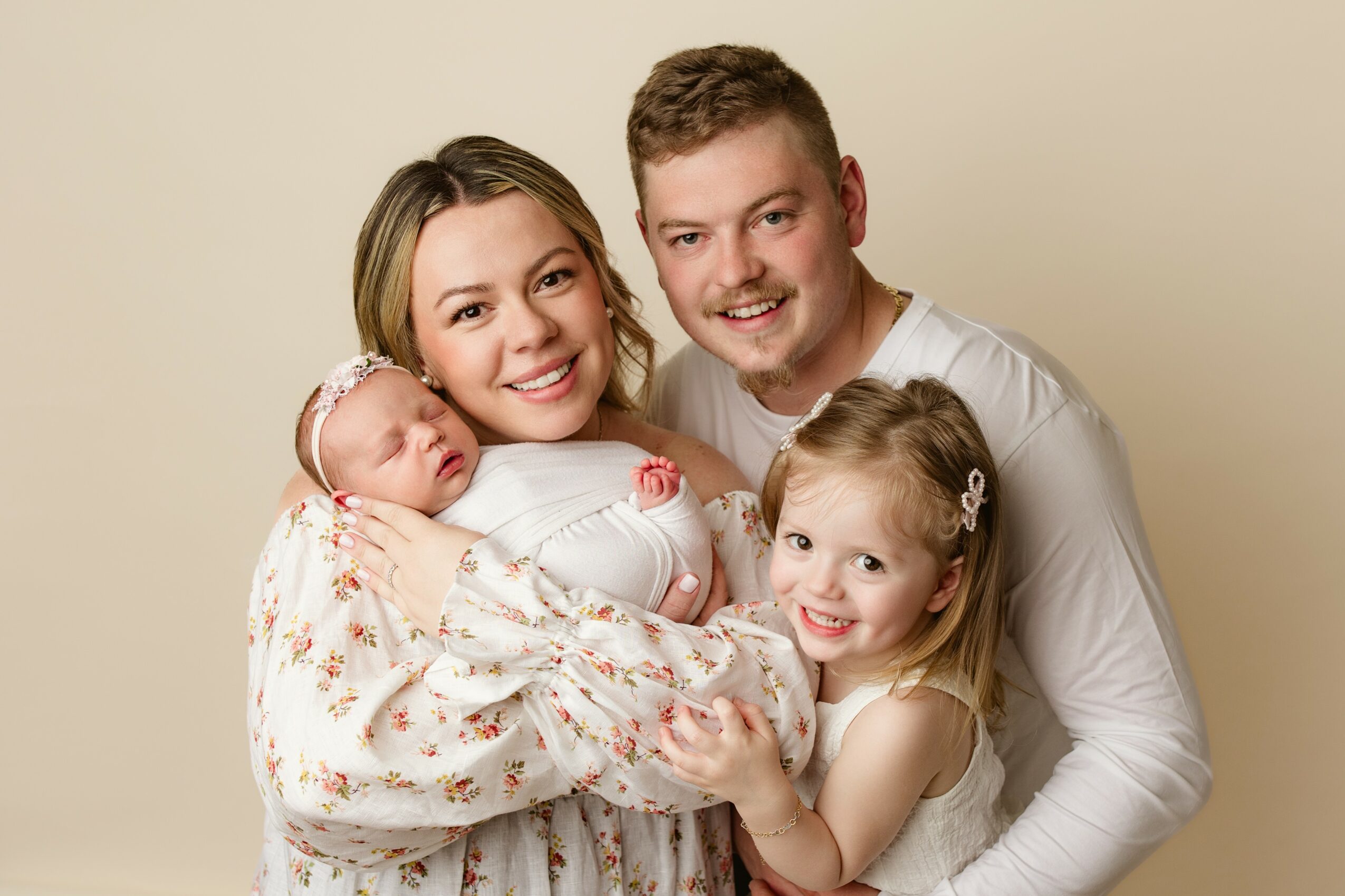 Family of four smiling, woman holding a newborn, man and young girl close together, neutral background.