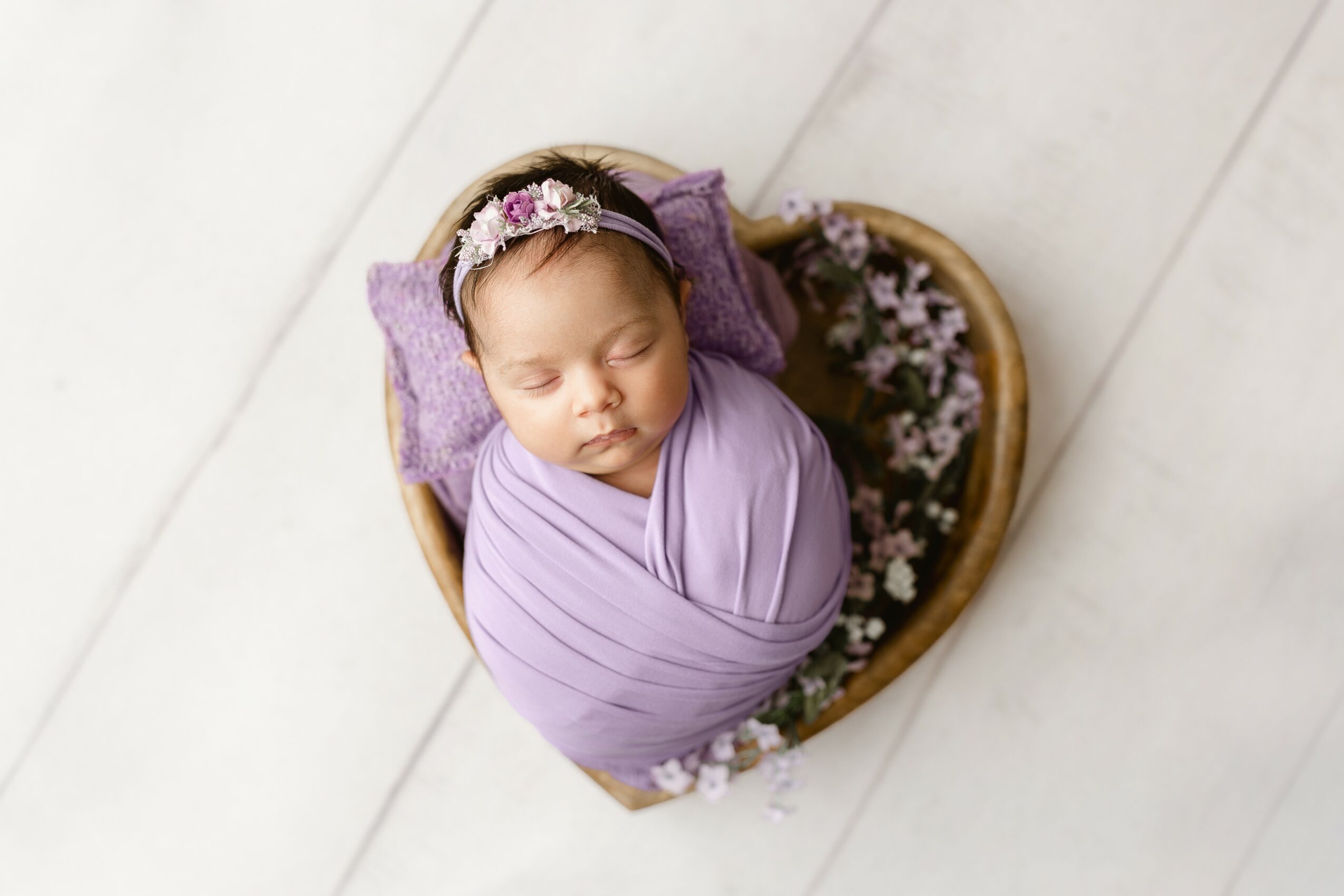 Baby girl wrapped in purple swaddle, sleeping in a basket with flowers, on a white floor.