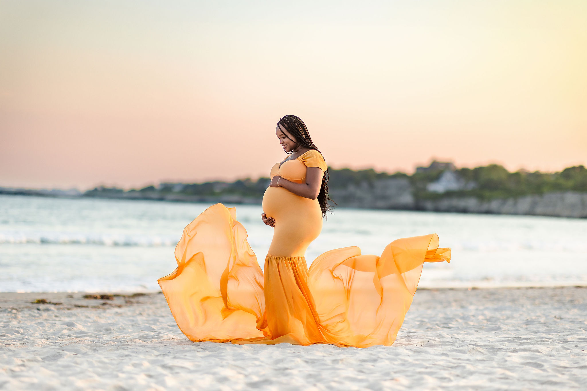 Pregnant woman in a flowing orange dress standing on a beach at sunset, with water and distant shoreline in background.
