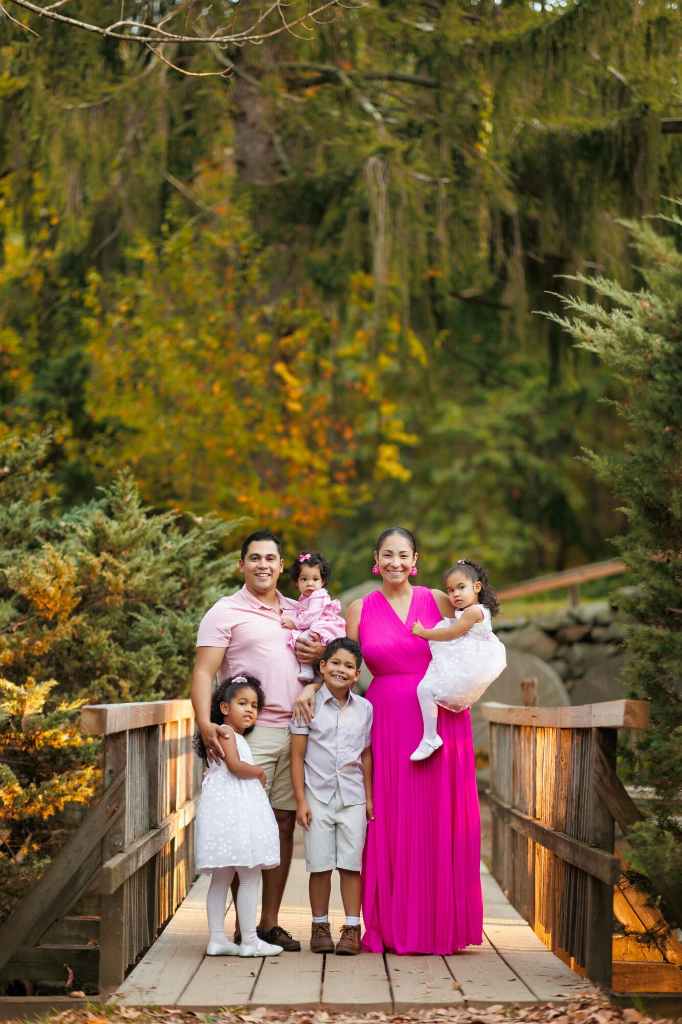Family of six standing on a wooden bridge surrounded by trees and foliage, smiling at the camera.