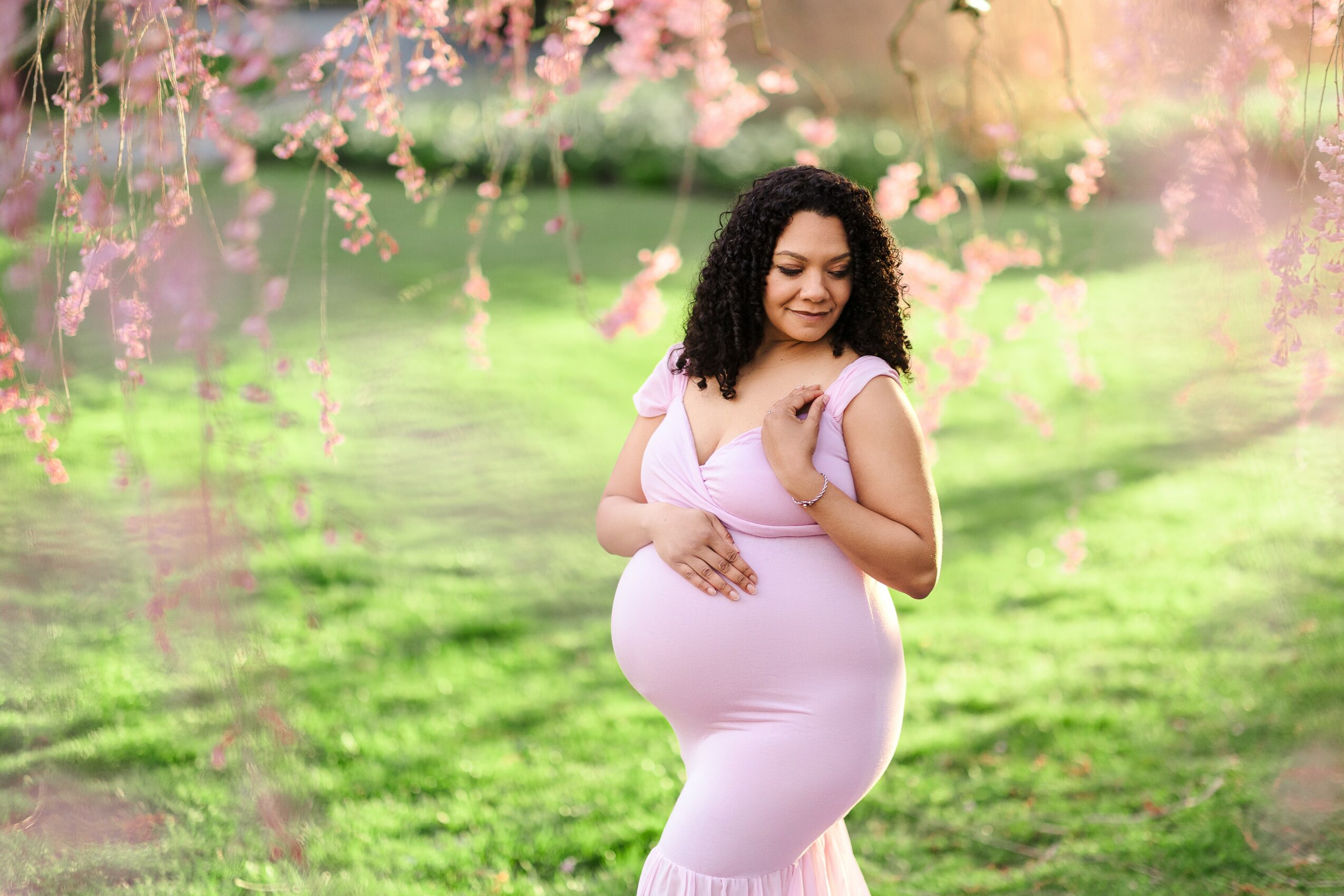 Pregnant woman with curly hair standing outdoors under pink flowering branches, smiling, with green grass background.