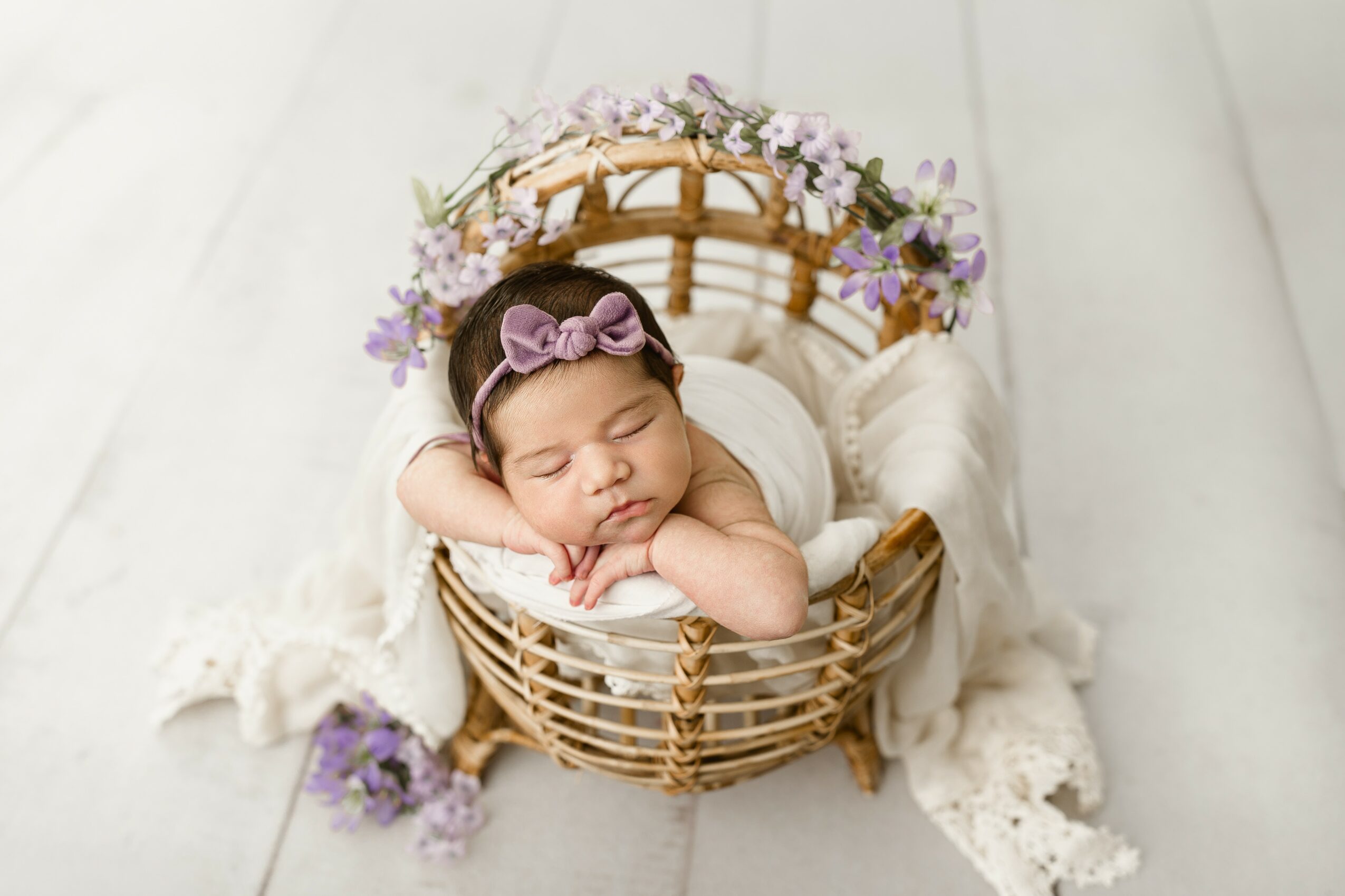 Baby girl with a pink bow headband sleeping in a wicker basket decorated with flowers and soft fabric, on a white surface.