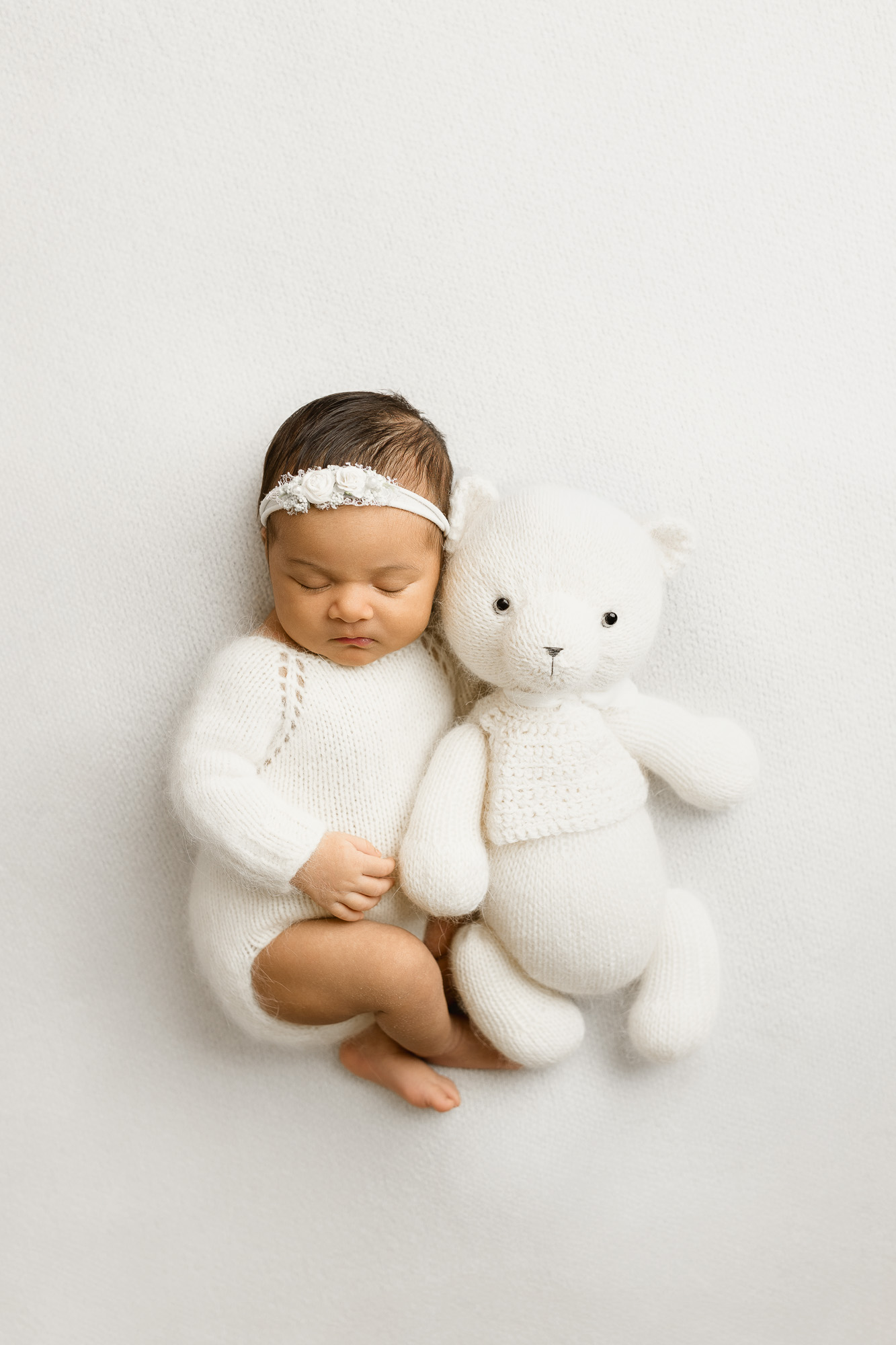 Baby girl with a headband, sitting next to a white teddy bear on a white background.