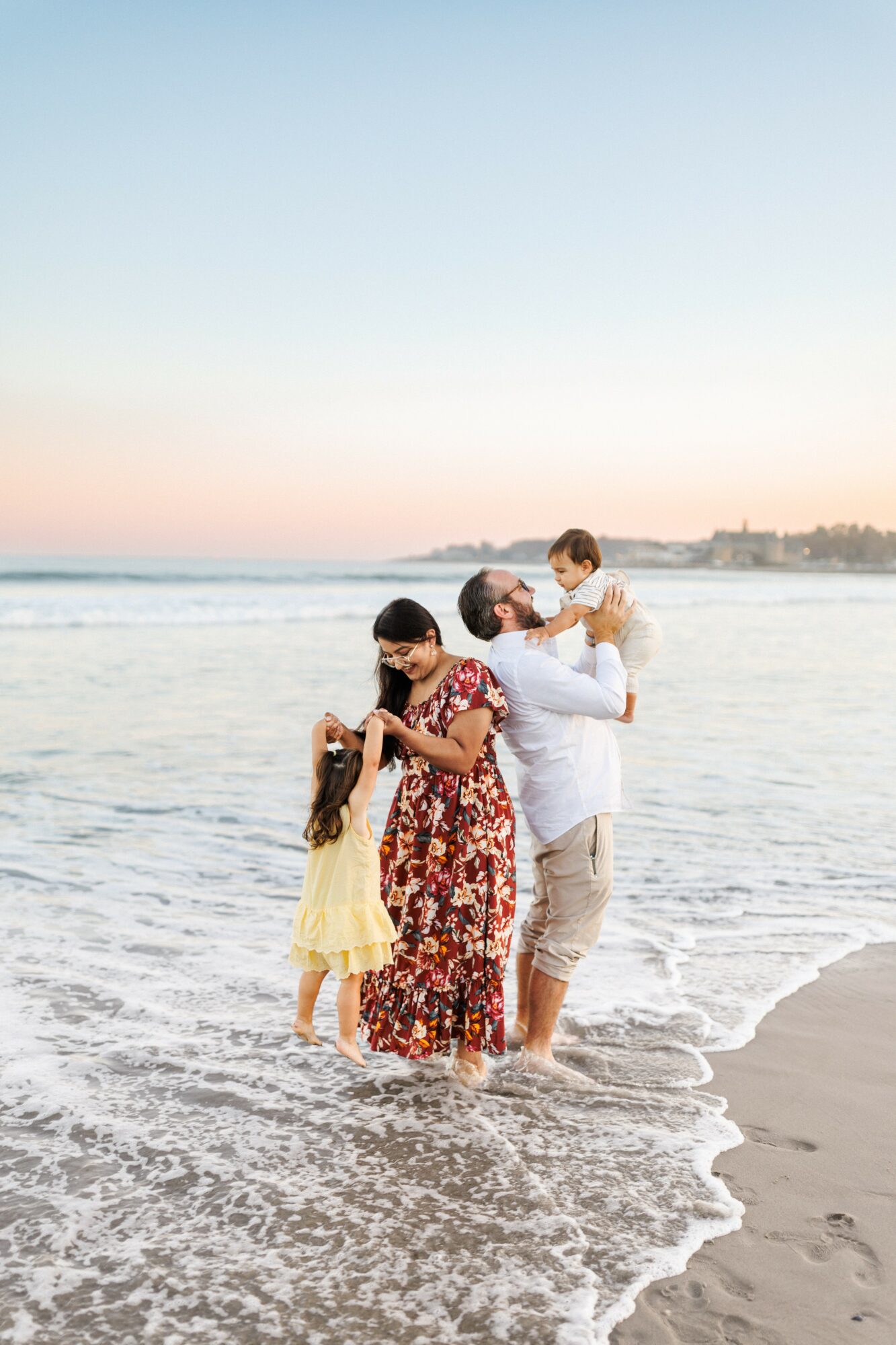 Family of four standing in shallow ocean water at sunset, with a beach and distant shoreline in background.