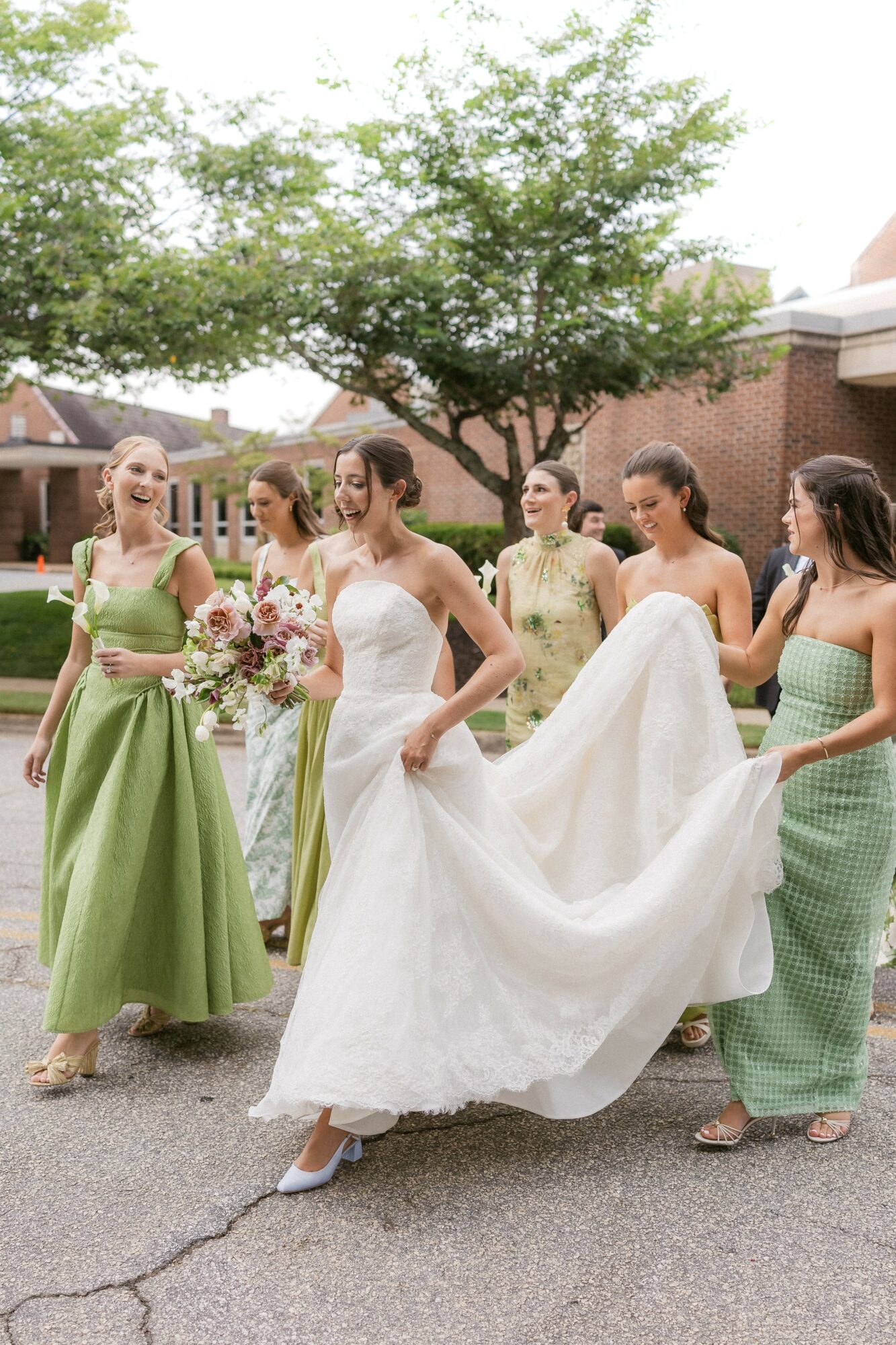 Bride in white dress with five women outdoors, one holding a bouquet, trees and buildings in background.