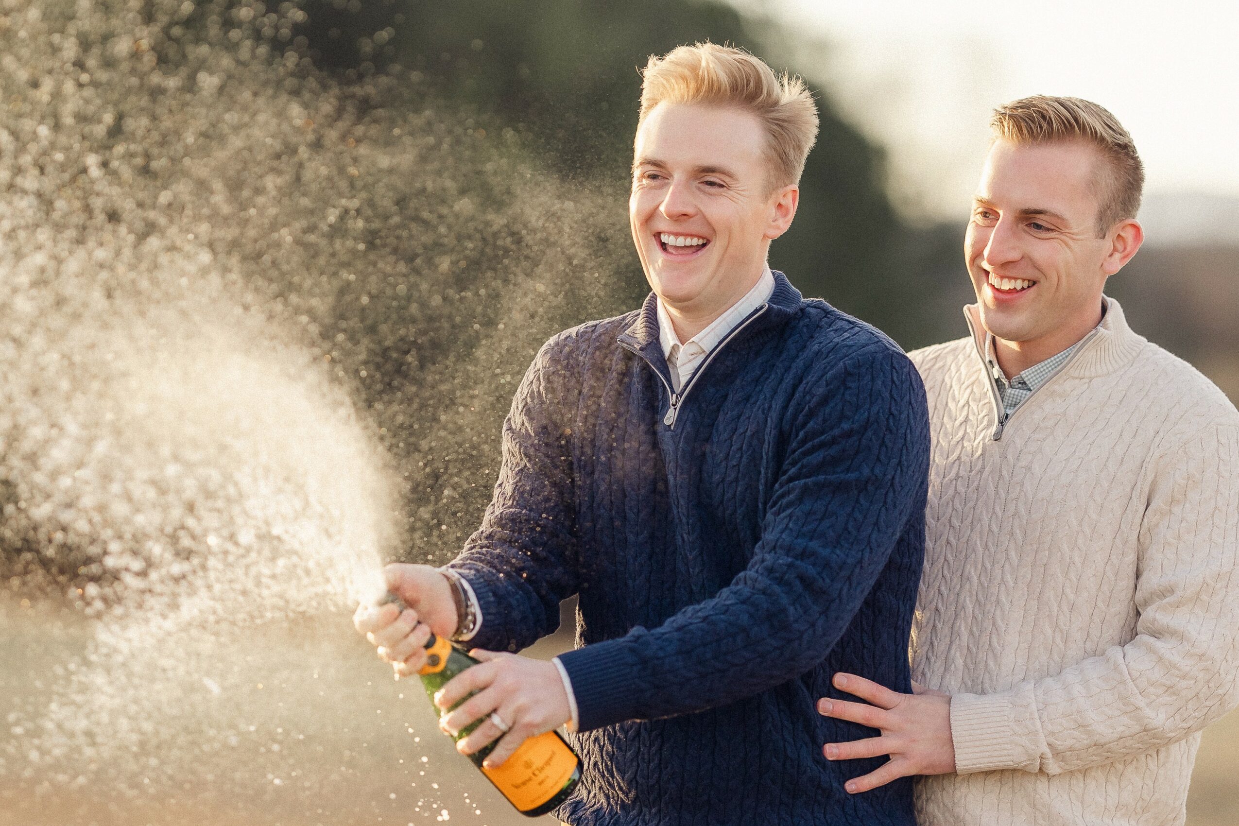 Two smiling men outdoors, one opening a champagne bottle with spray, the other touching his back.