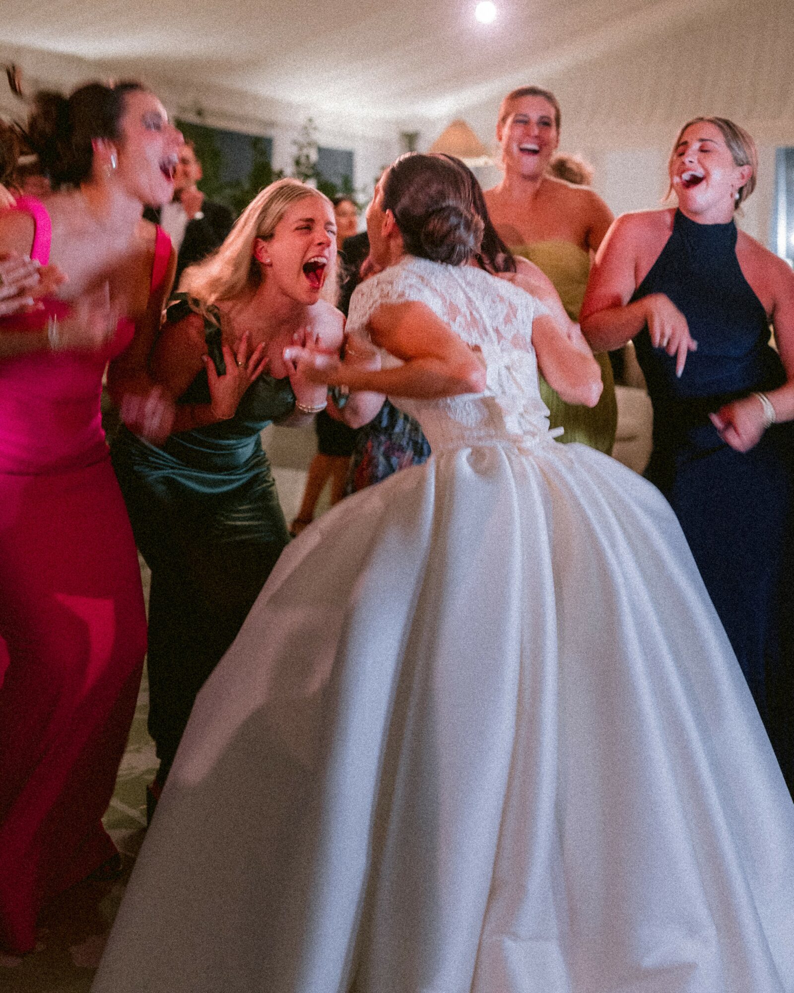 Group of women in formal dresses laughing and celebrating around a bride in a wedding gown indoors.