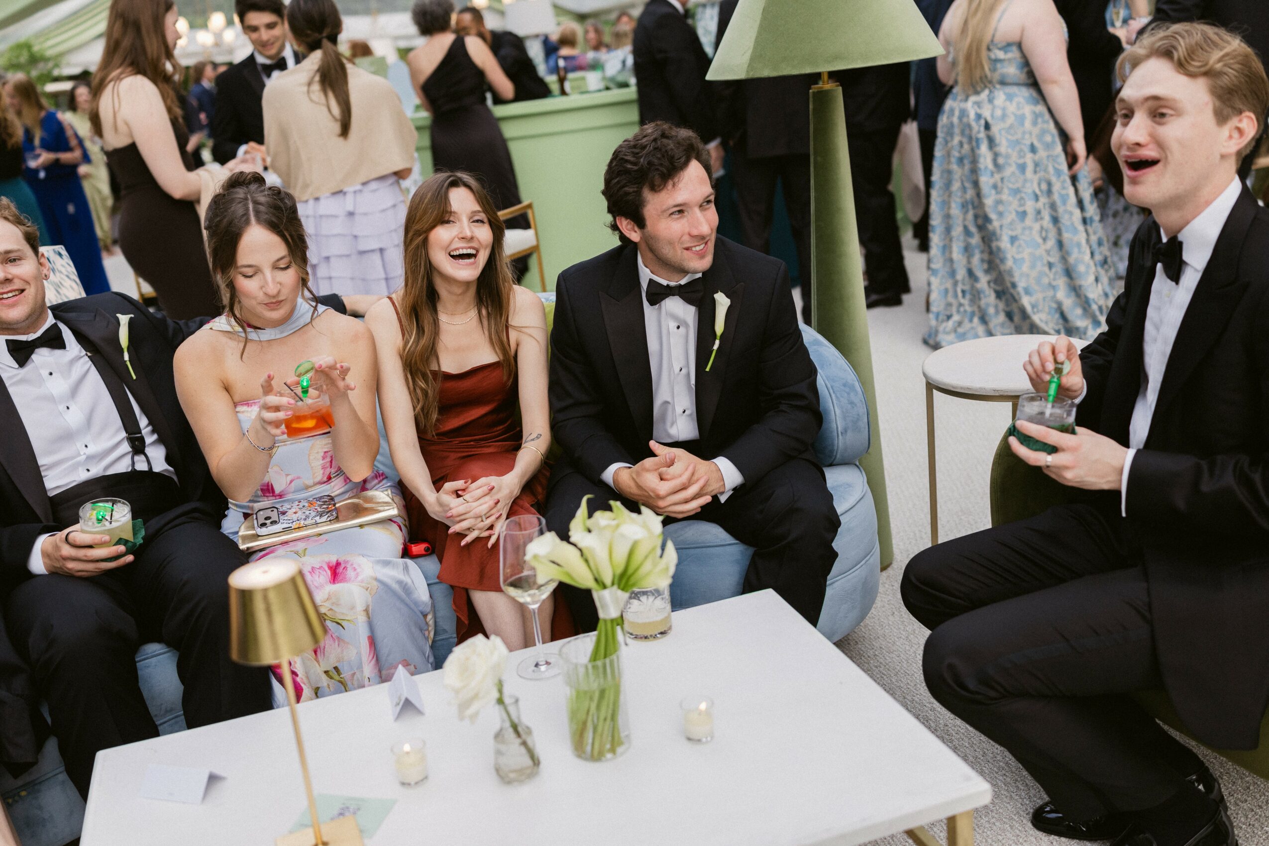 Group of people in formal attire sitting and standing around a table with drinks and flowers at a social event.