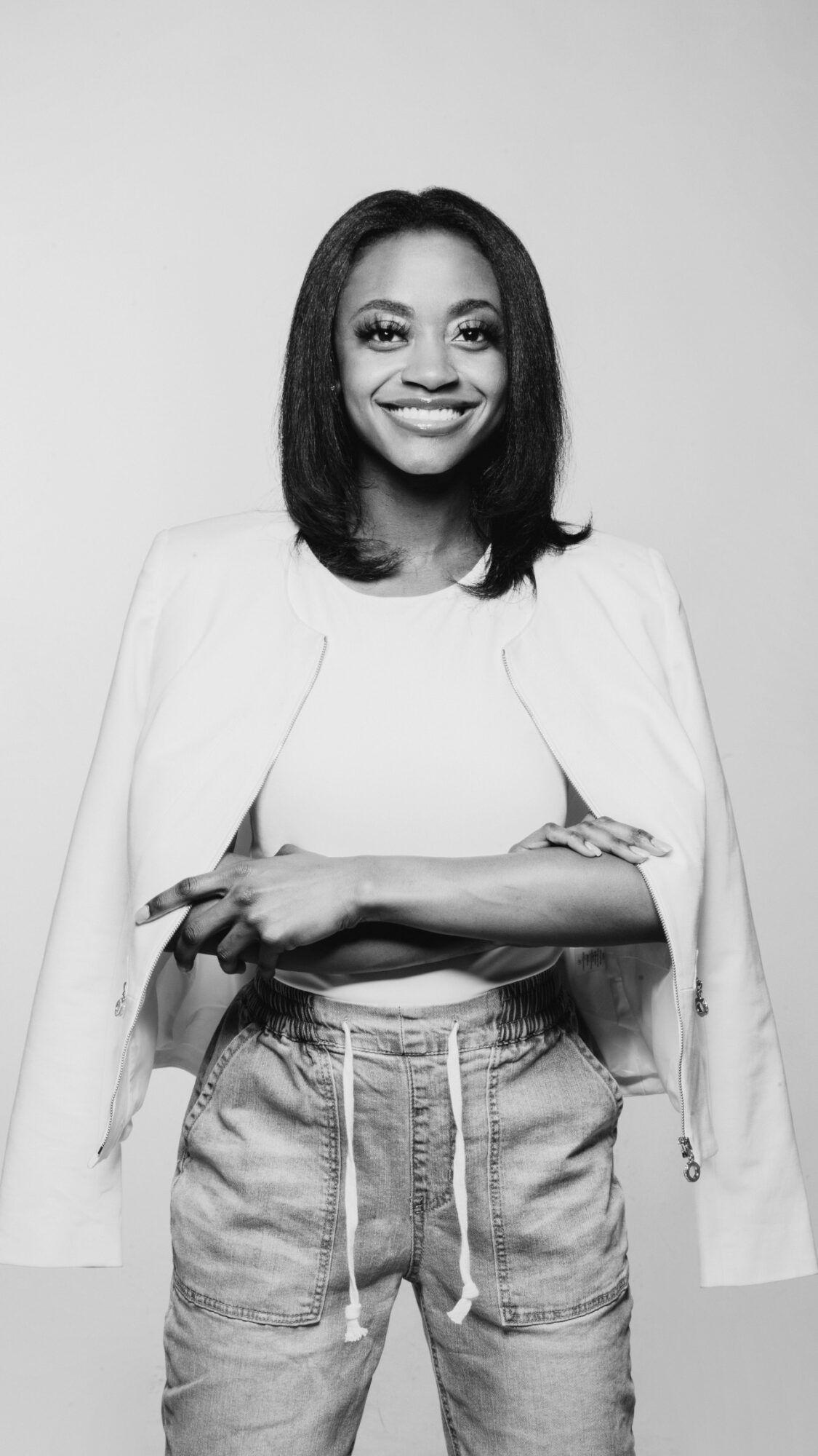 Young woman smiling, wearing a white jacket over a top and jeans, with arms crossed, in black and white.