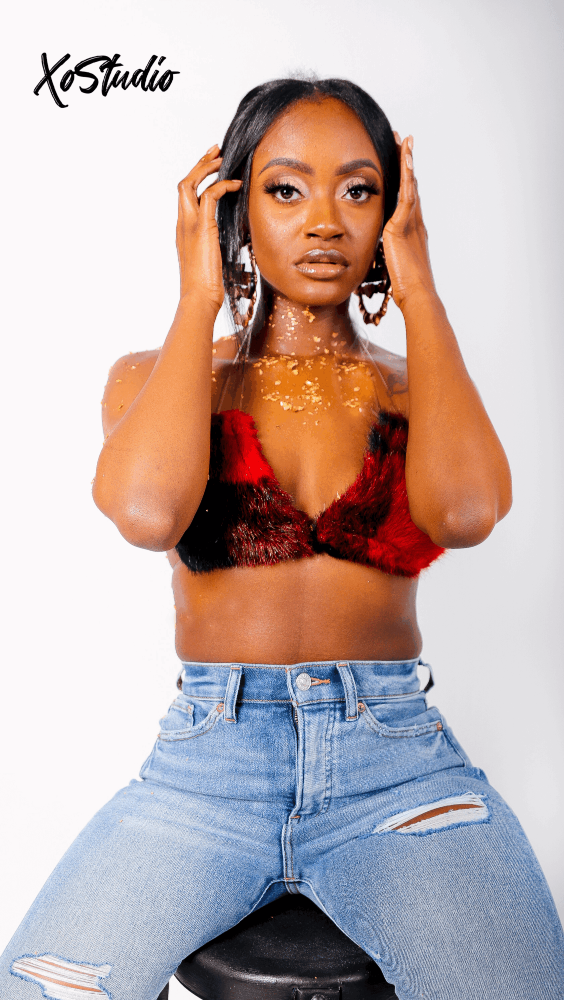 Young woman with long dark hair, wearing a red and black top, jeans, and jewelry, posing with hands near her face.