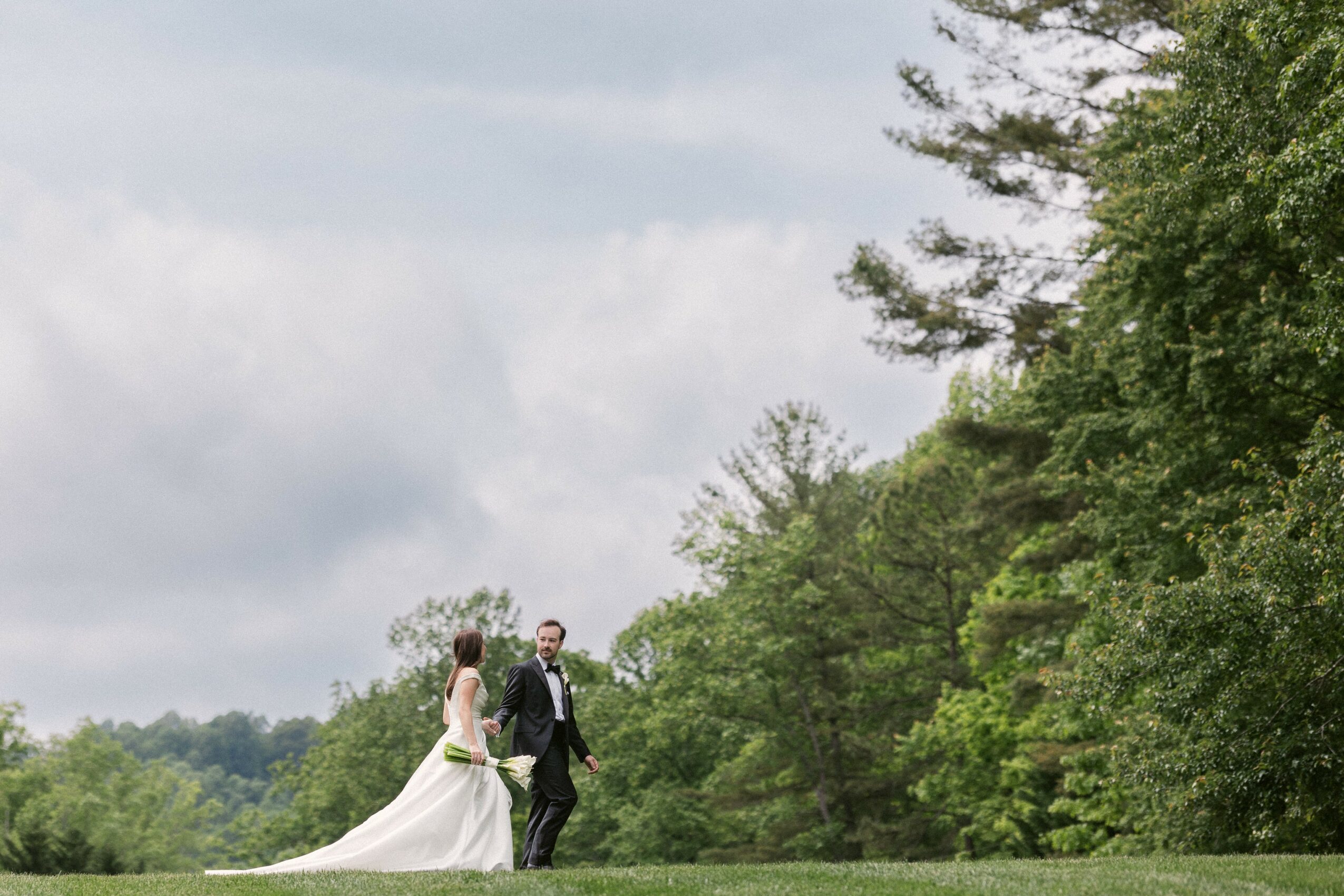 A bride and groom walking hand in hand outdoors with trees and cloudy sky in background.