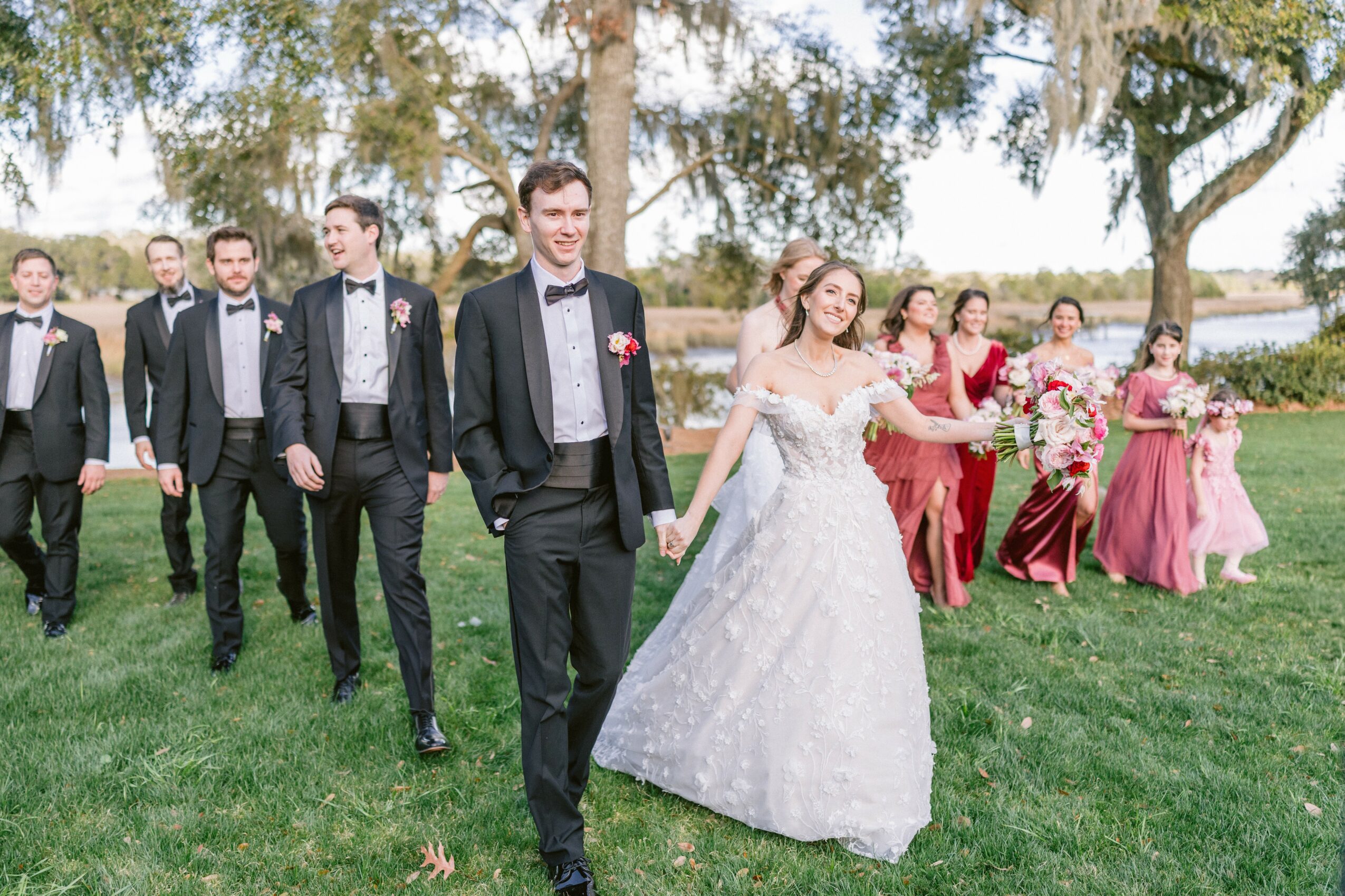 Bride and groom walking outdoors with wedding party, trees, and water in background, smiling and holding hands.