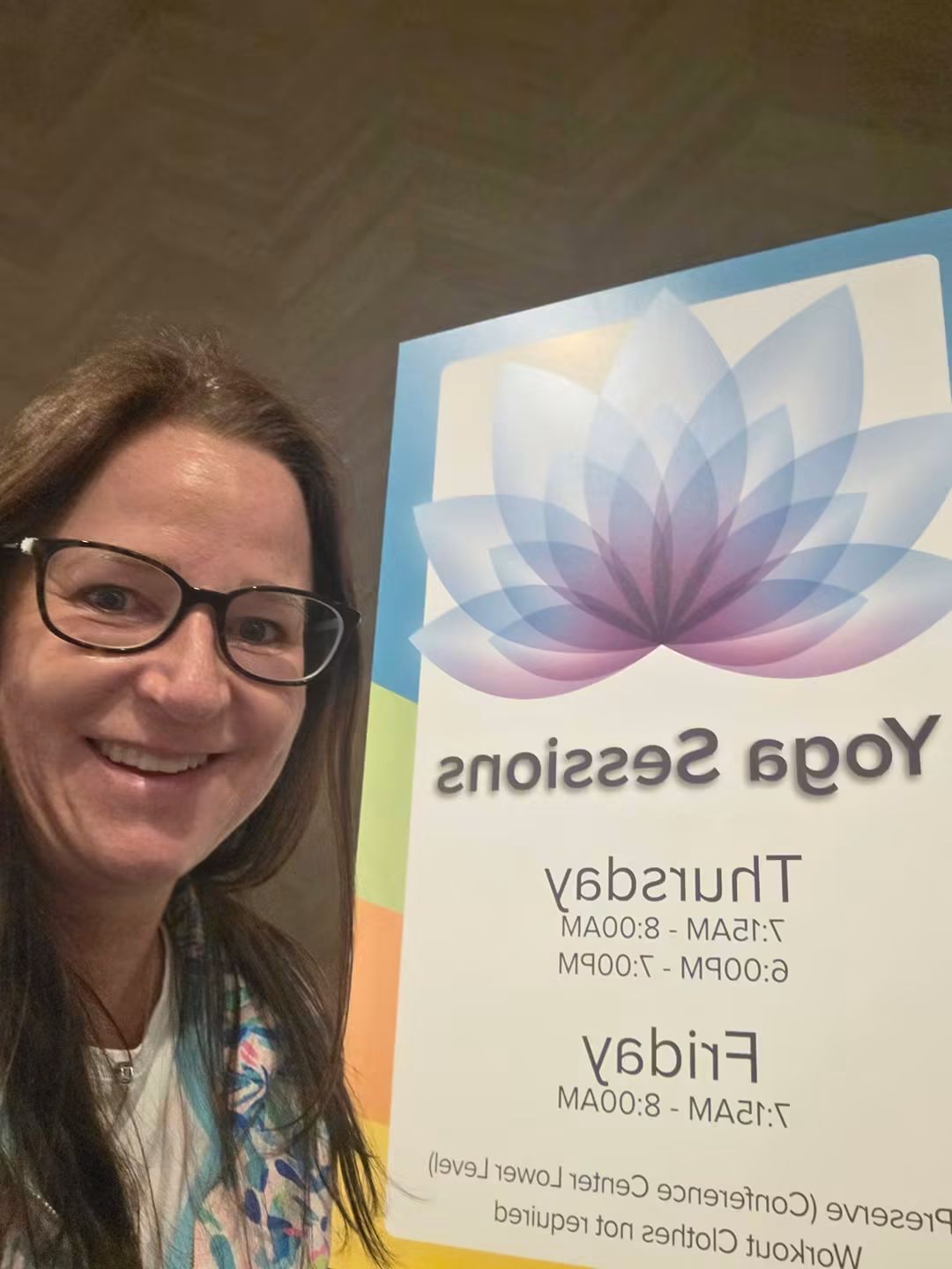 Woman with glasses smiling next to a colorful sign with a lotus flower design, text about yoga sessions, indoors.