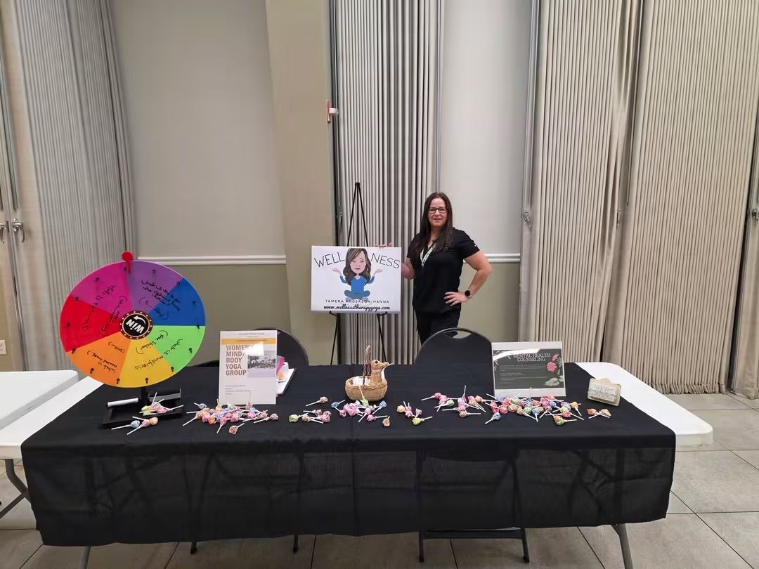 Woman standing behind a table with promotional items, a colorful wheel, and a sign, in a room with vertical blinds.