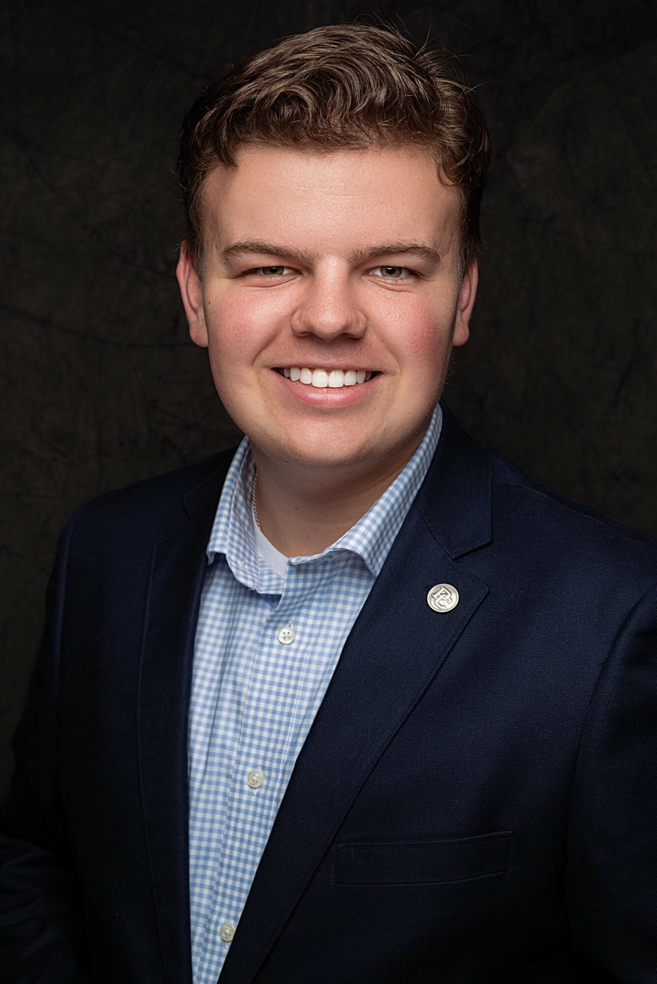 Young man with short hair smiling, wearing a dark blazer and light blue checkered shirt, against a dark background.
