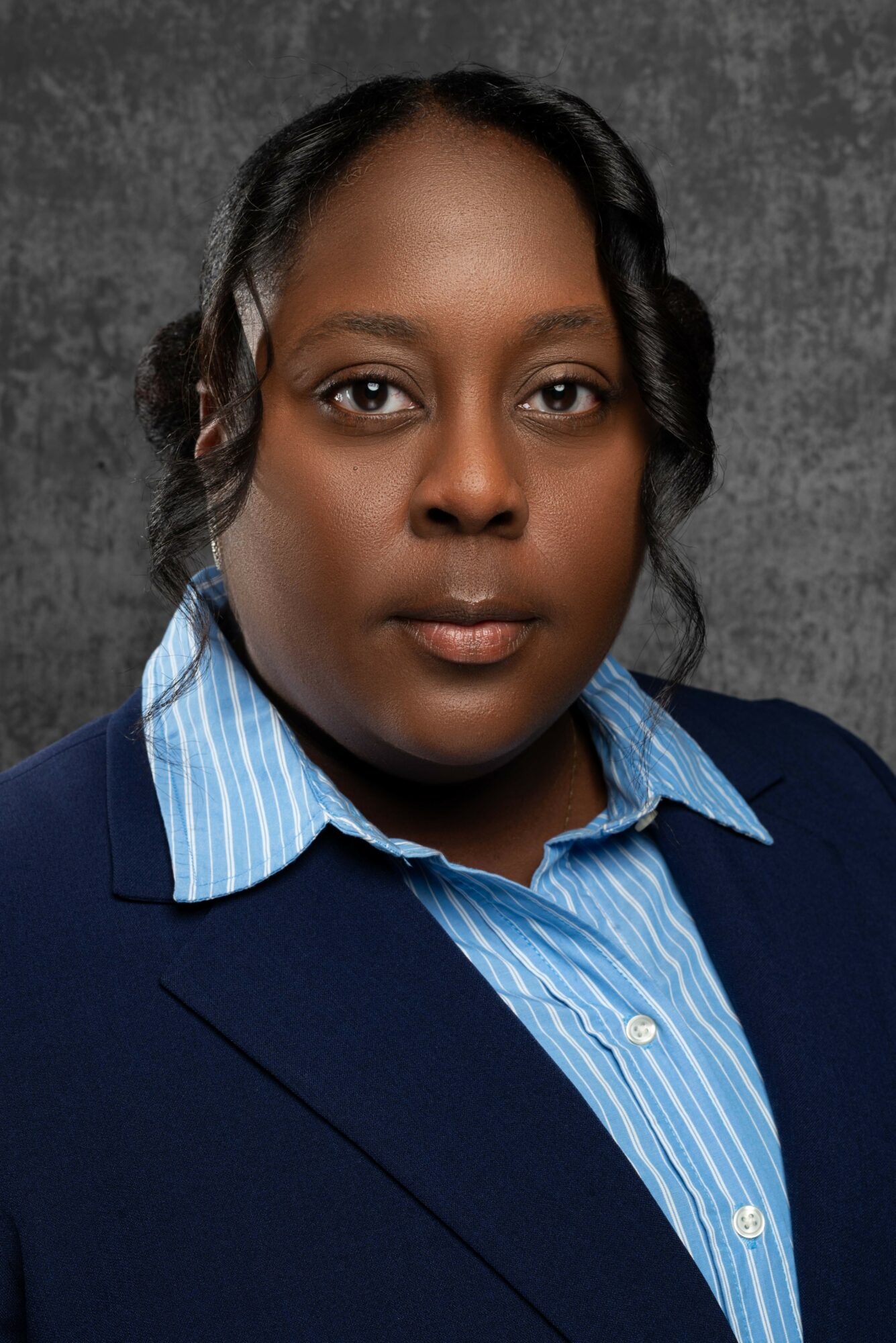 Portrait of a woman with dark hair, wearing a blue striped shirt and blazer, against a dark textured background.