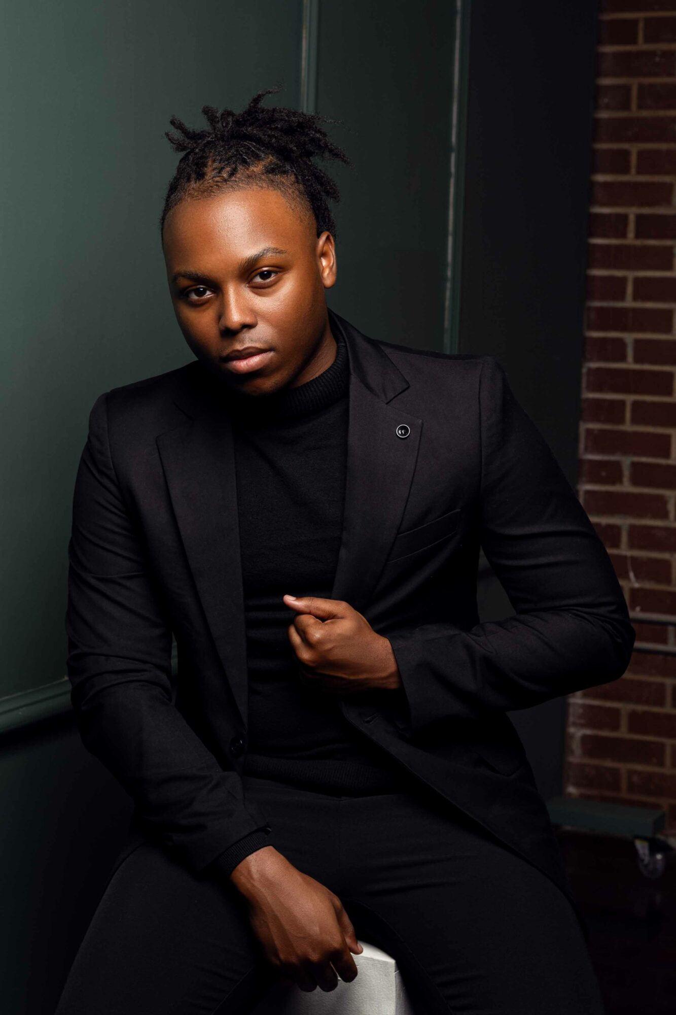 Young person with dreadlocks wearing a black blazer and shirt, standing against a dark background with brick wall.