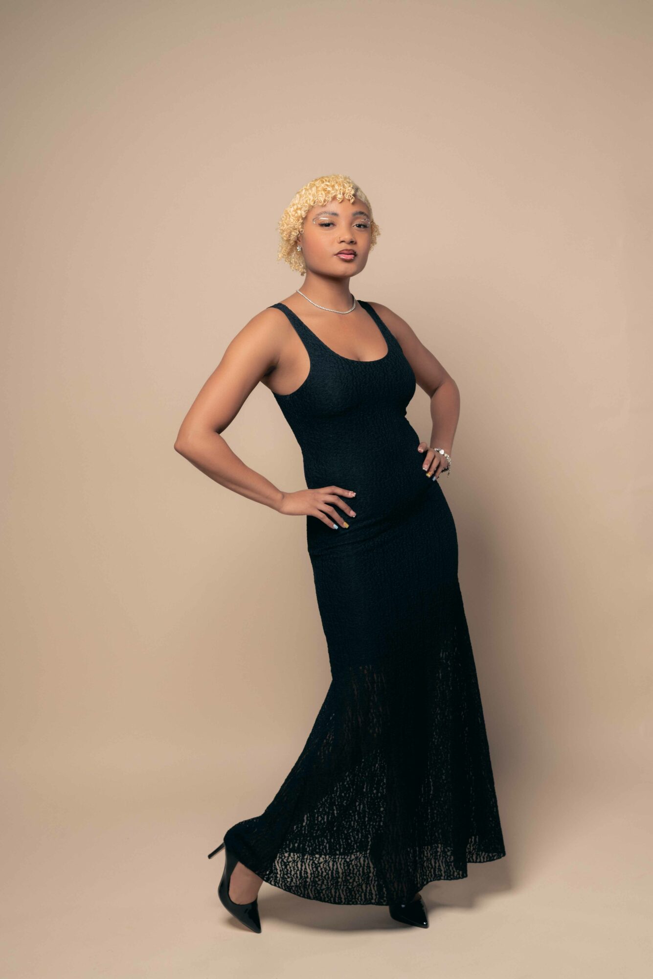 Woman with short curly blonde hair in a black sleeveless dress and high heels, standing with hands on hips against a plain background.