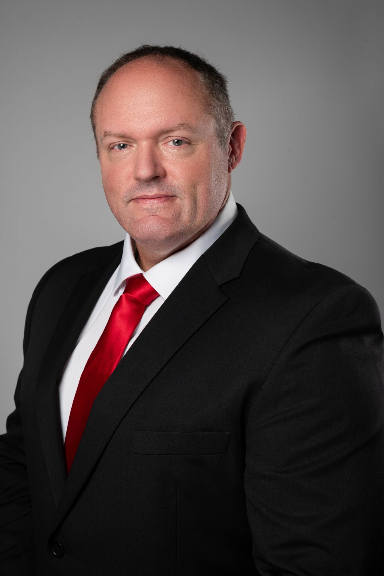 Man in a black suit, white shirt, and red tie poses against a gray background.