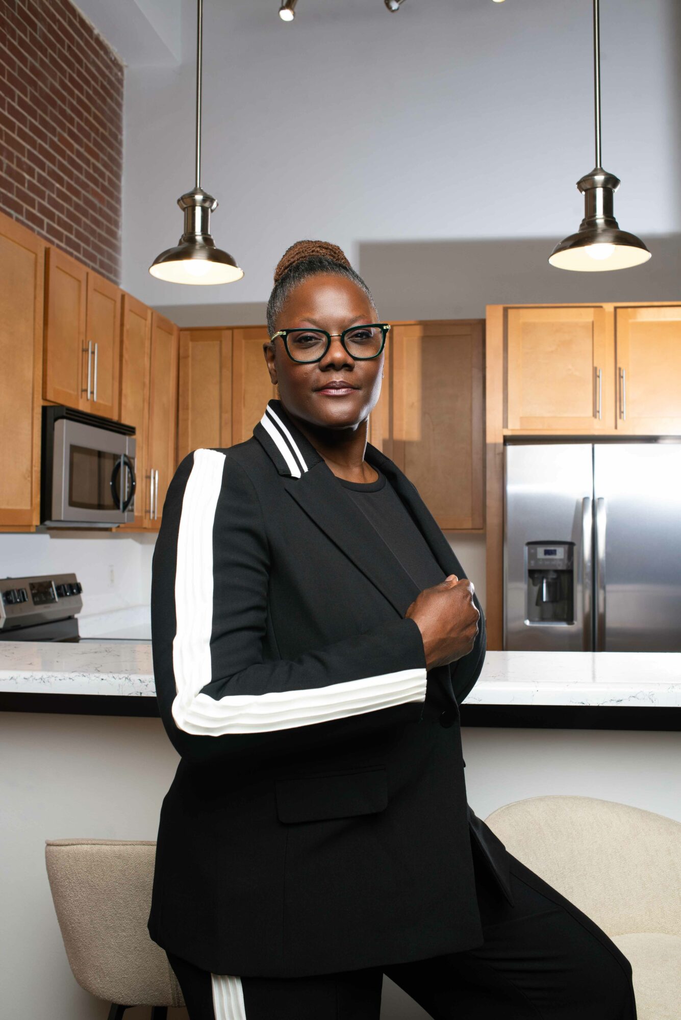 Woman with glasses and a black jacket with white stripes on sleeves standing in a kitchen.