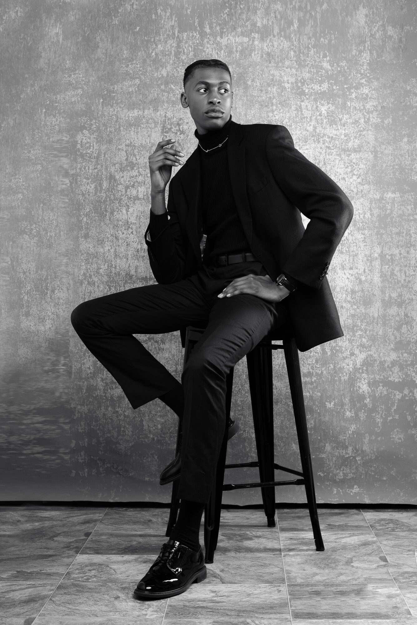 Person sitting on a stool, wearing a dark suit and holding a cigarette, against a textured background.