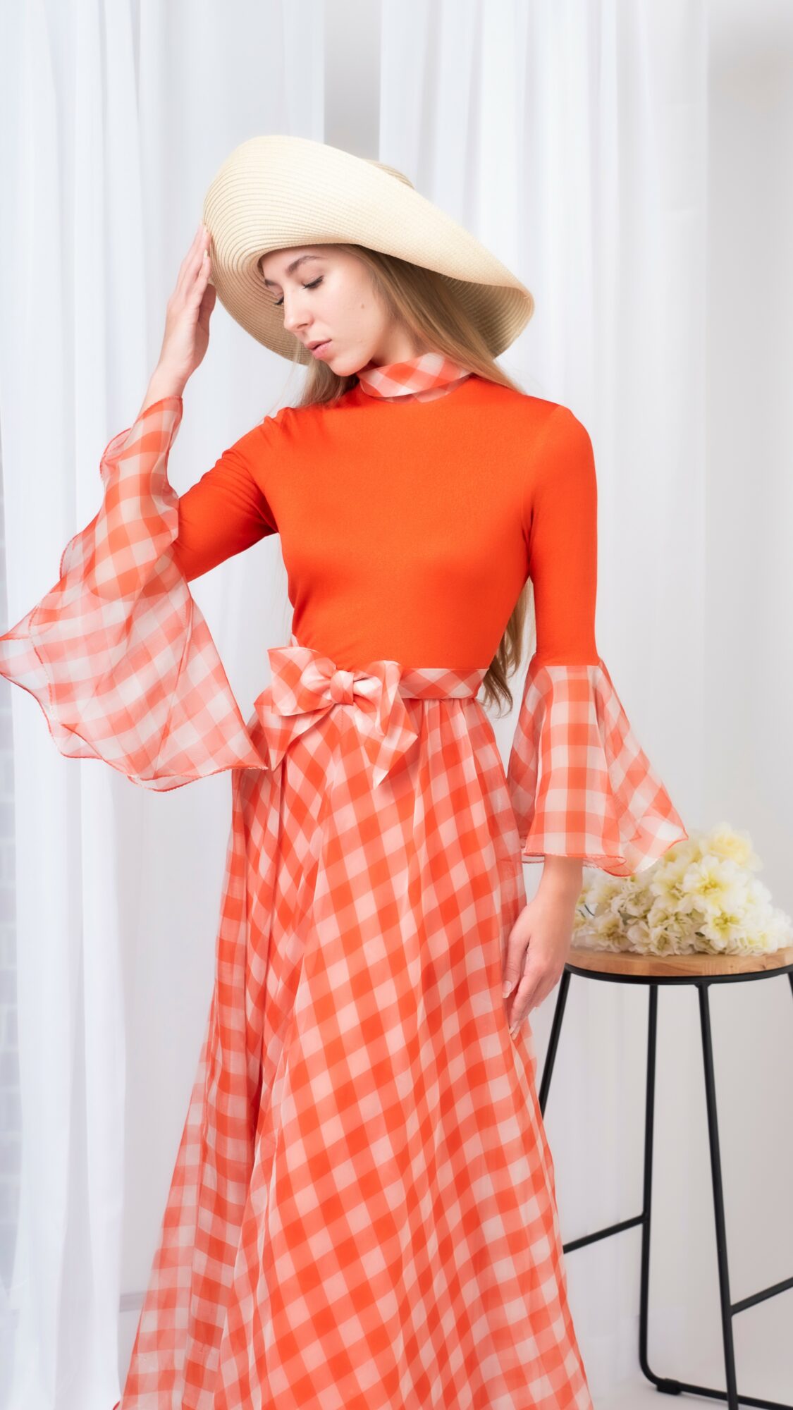 Woman in orange dress with checkered ruffled sleeves and skirt, wearing a wide-brimmed hat, standing near a small table with flowers.