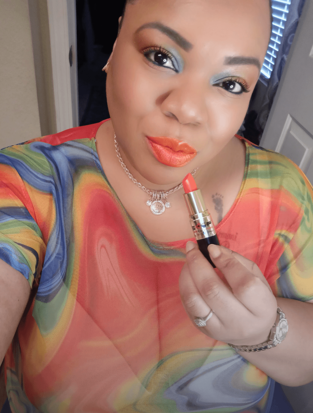 Woman with colorful top applying coral lipstick, wearing jewelry, in a room with a door and mirror behind her.