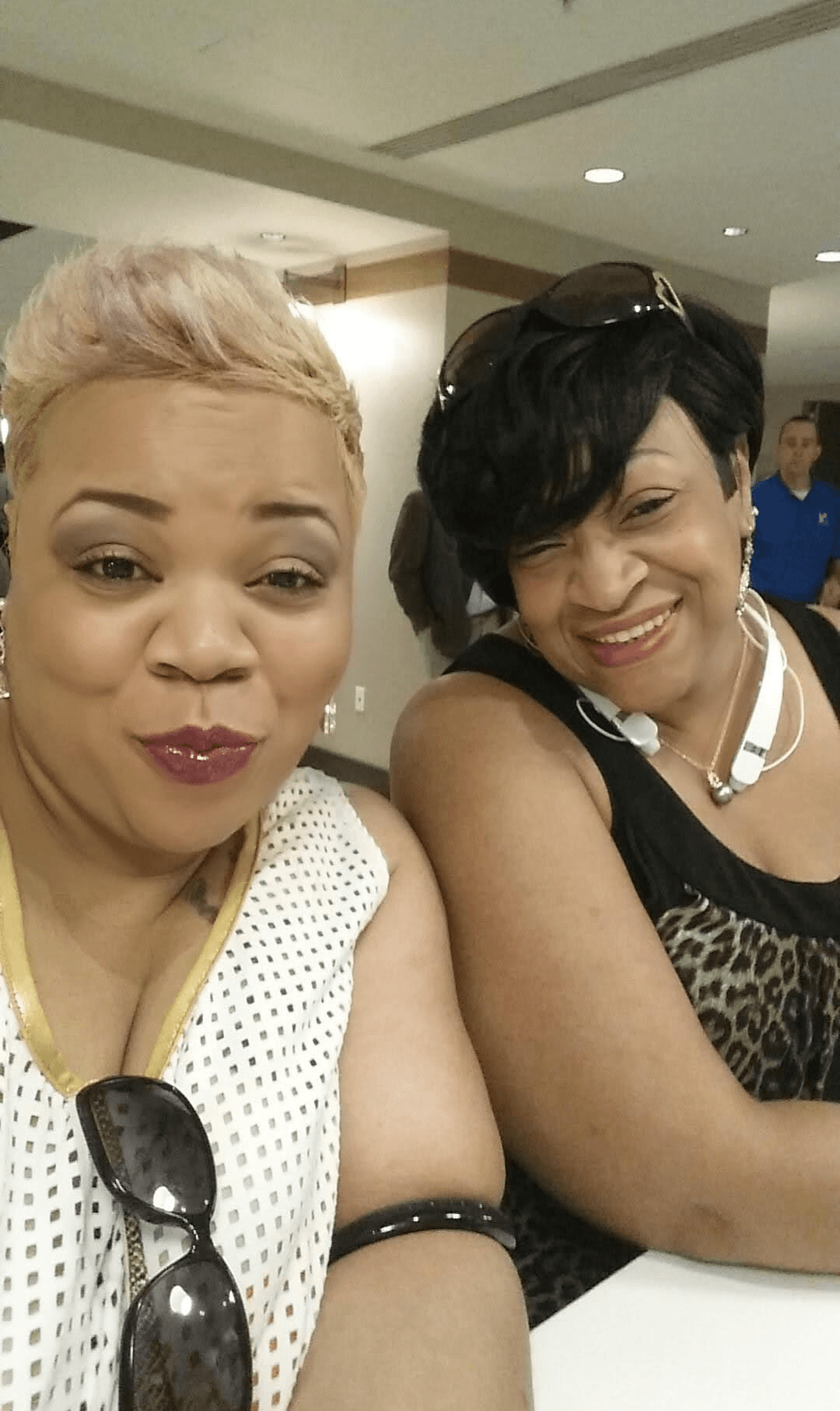 Two women smiling, one with short blonde hair and the other with short black hair, sitting indoors.