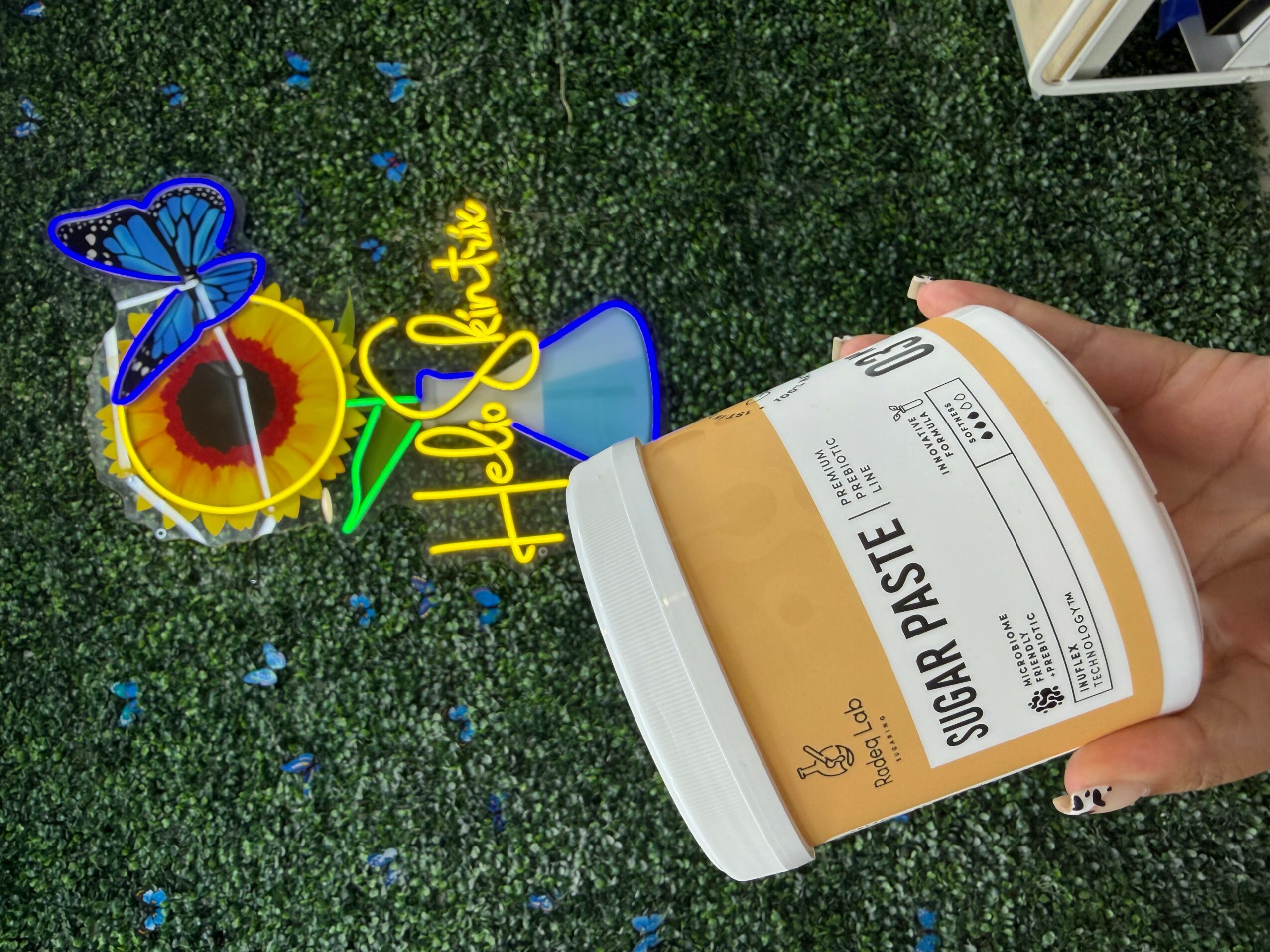 Hand holding a container of sugar paste with a green, blue, and yellow decorative sign on a green textured surface.