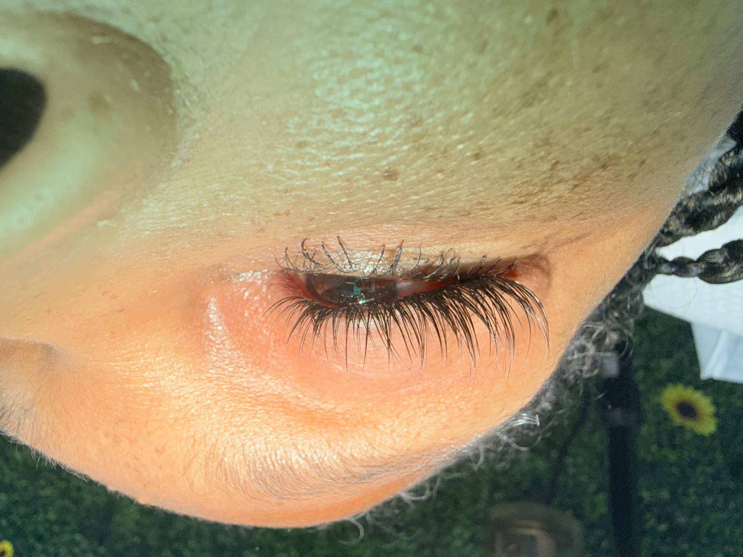 Close-up of a person's eye with long eyelashes, closed, showing skin texture and part of the nose, with a blurred background.