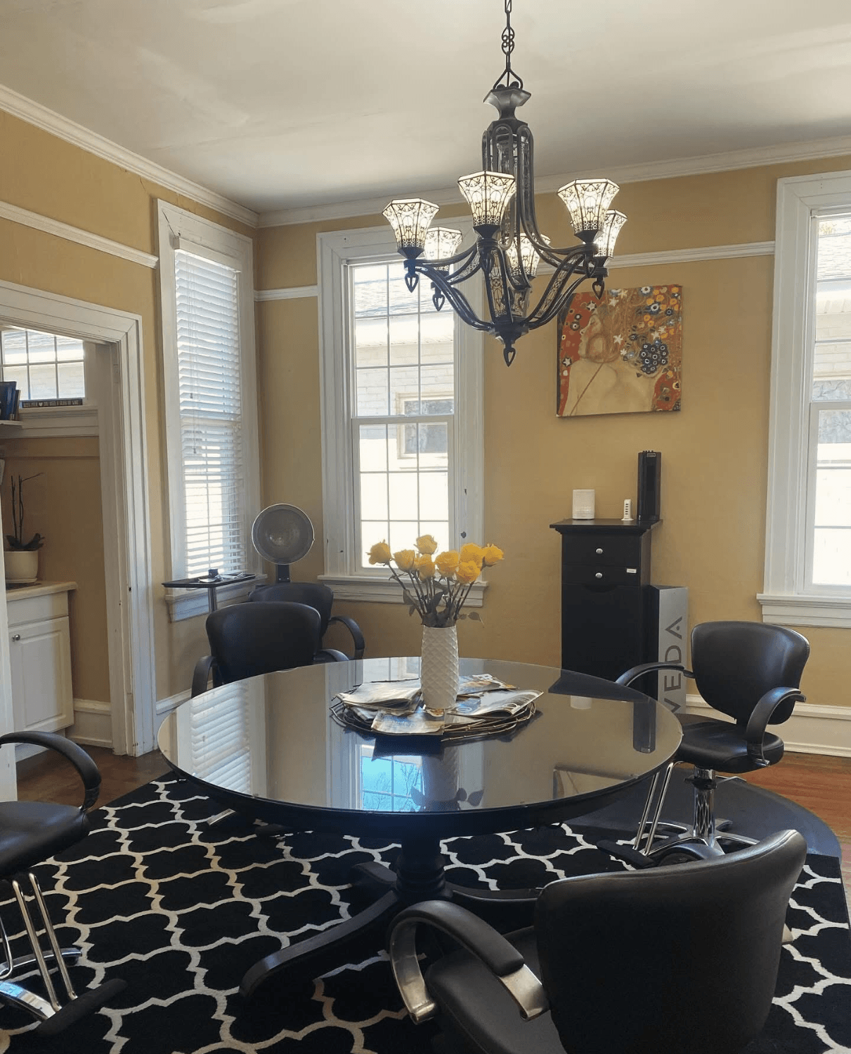 Dining room with round glass table, black chairs, chandelier, and windows with blinds, beige walls, and a black and white rug.