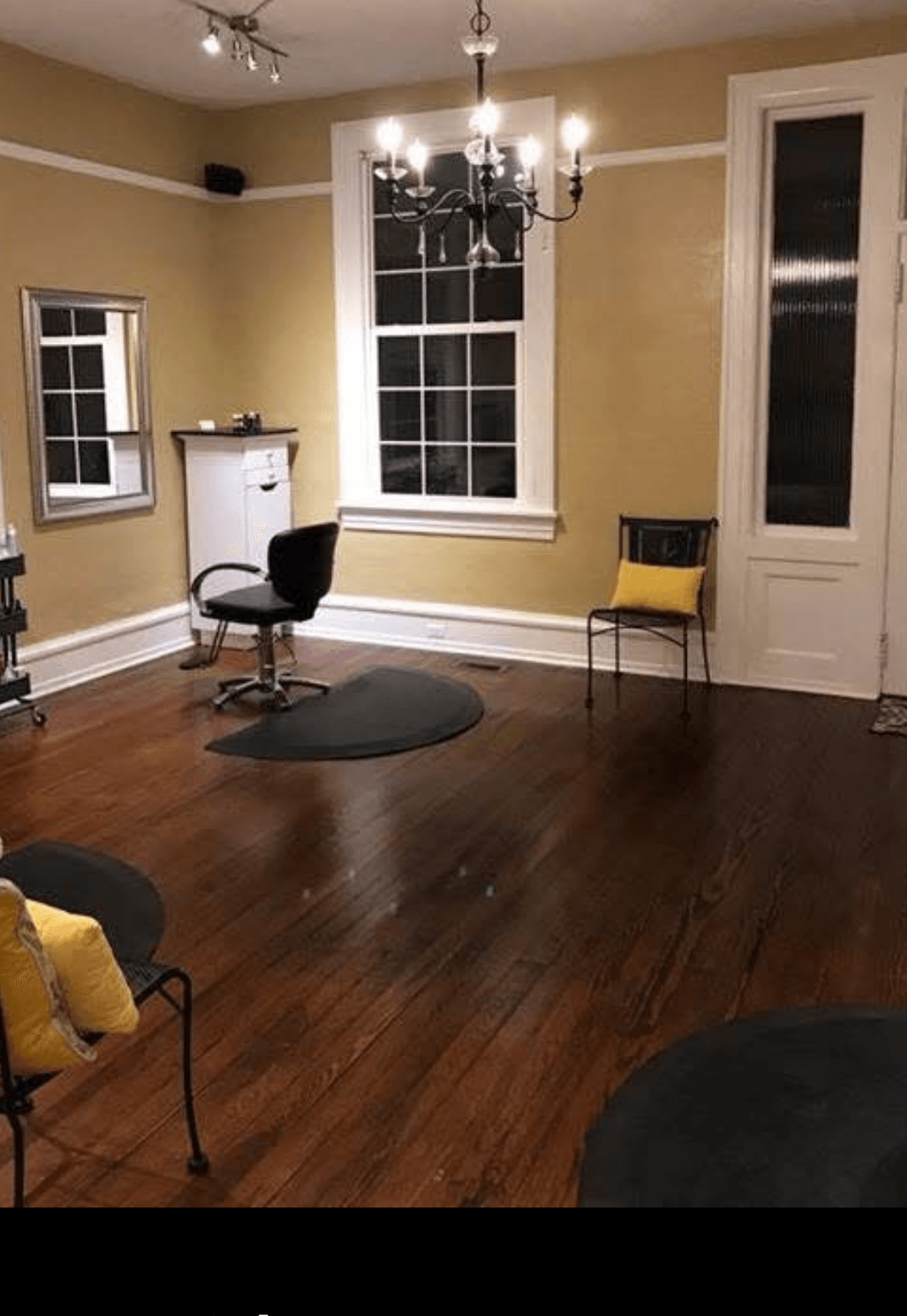 Room with wooden floor, two large windows, a desk with a chair, and a chandelier hanging from the ceiling.