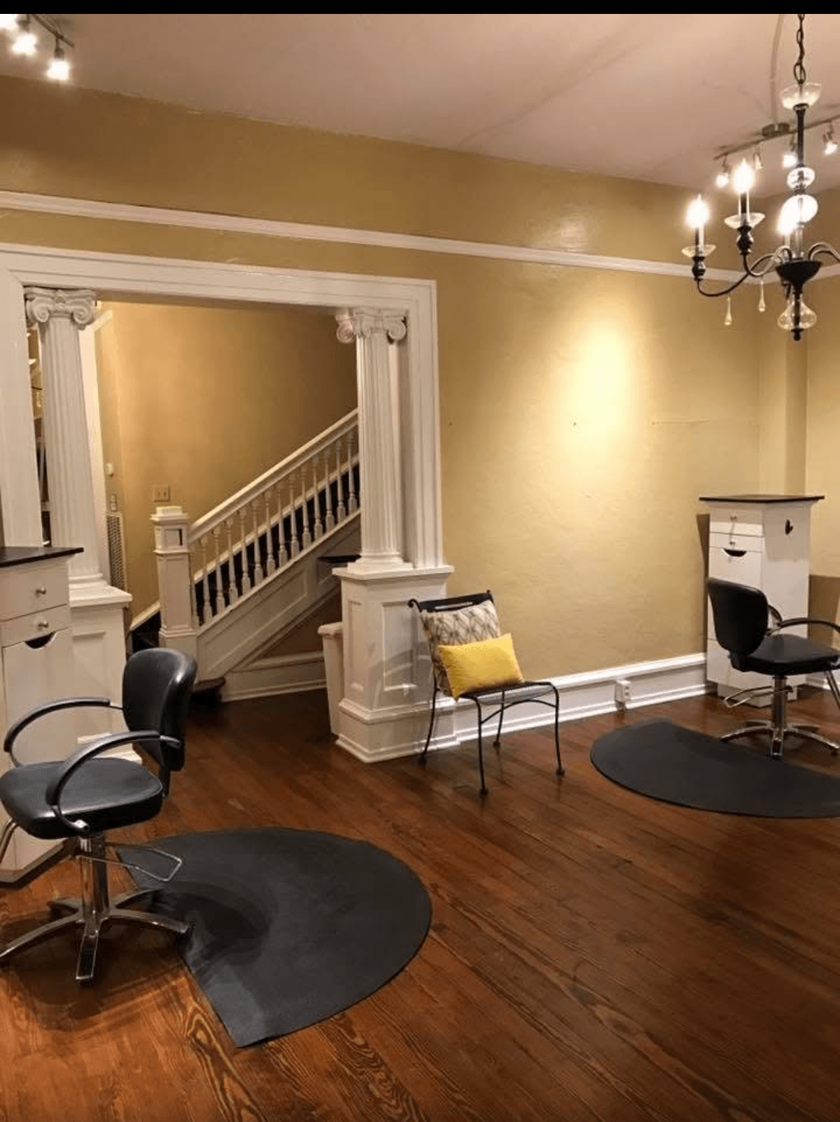 Room with wooden floor, chandelier, chairs, and a staircase in the background, beige walls, and decorative columns.