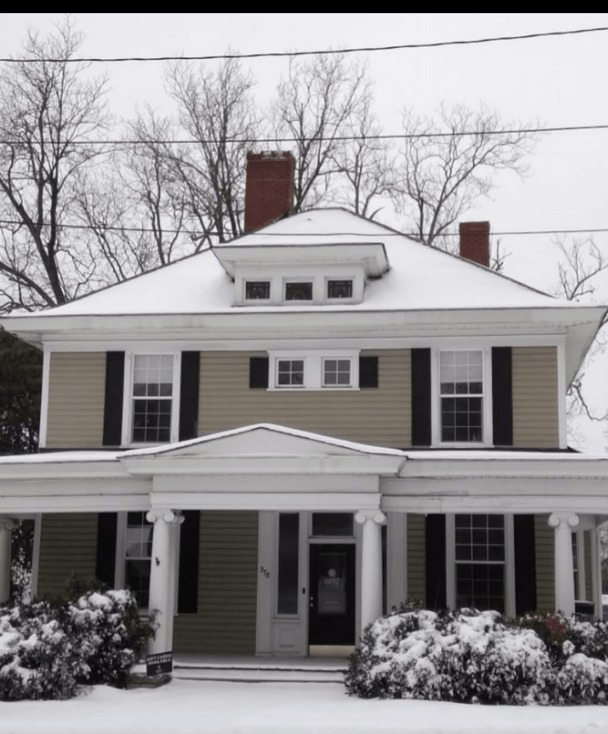 Two-story house with a porch, snow-covered bushes, and leafless trees in the background, overcast sky, and power lines above.