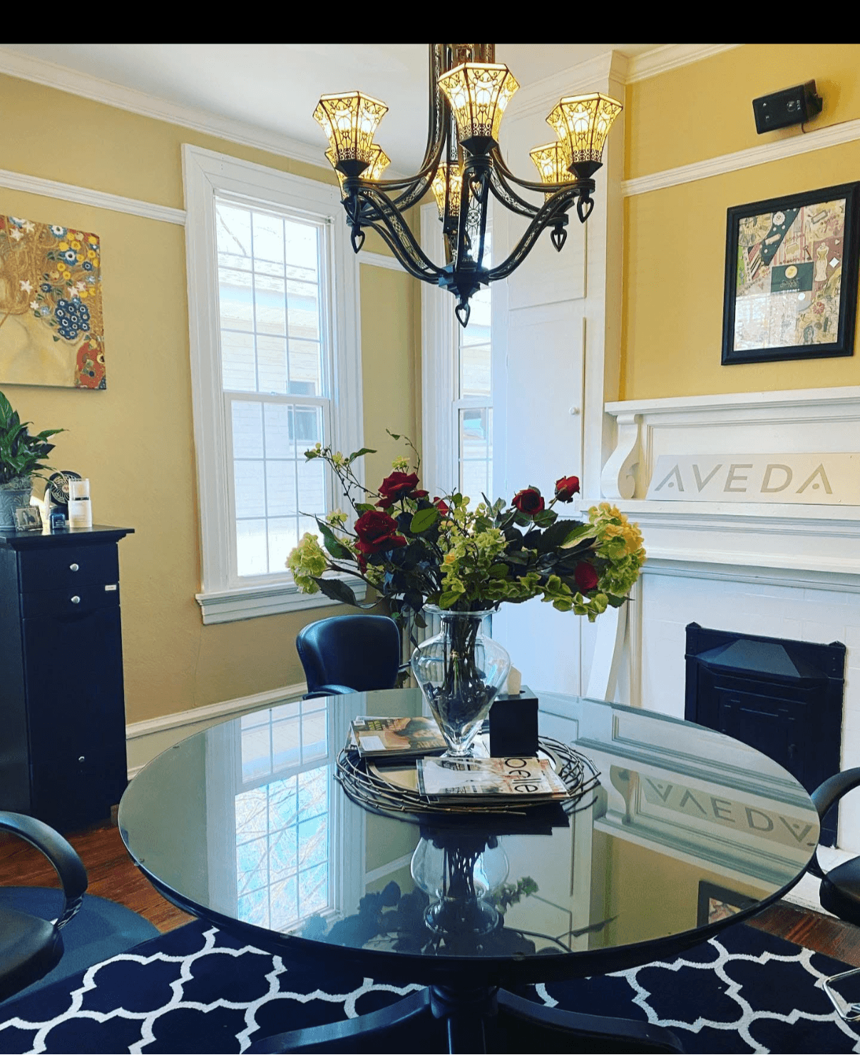 Round glass dining table with a large flower arrangement, chandelier overhead, and windows in a yellow-walled room.