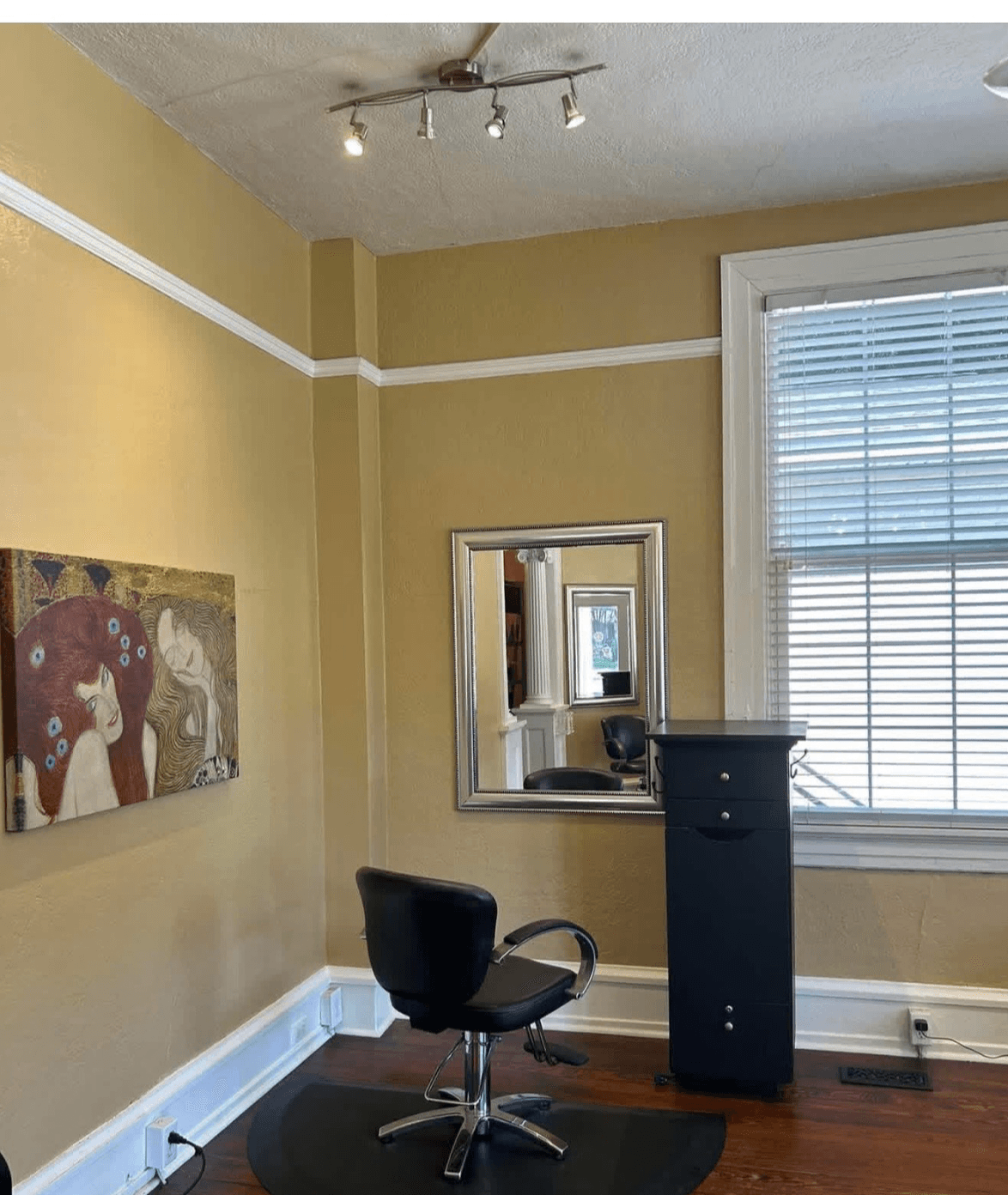 Corner of a room with a black chair, a mirror, a tall black cabinet, a window with blinds, and a painting on a yellow wall.